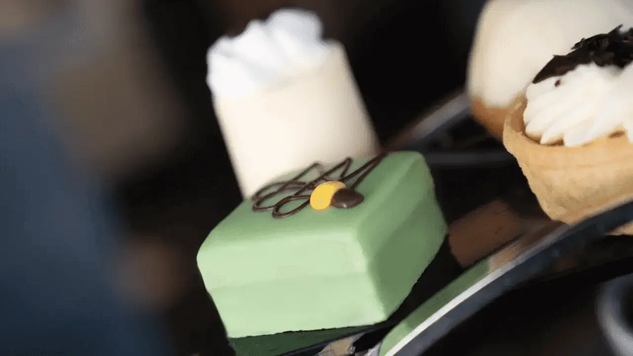 Close-up of assorted desserts including a green mousse cake with chocolate and yellow decoration, topped with whipped cream and chocolate shavings, and a small tart with whipped cream and chocolate shavings.