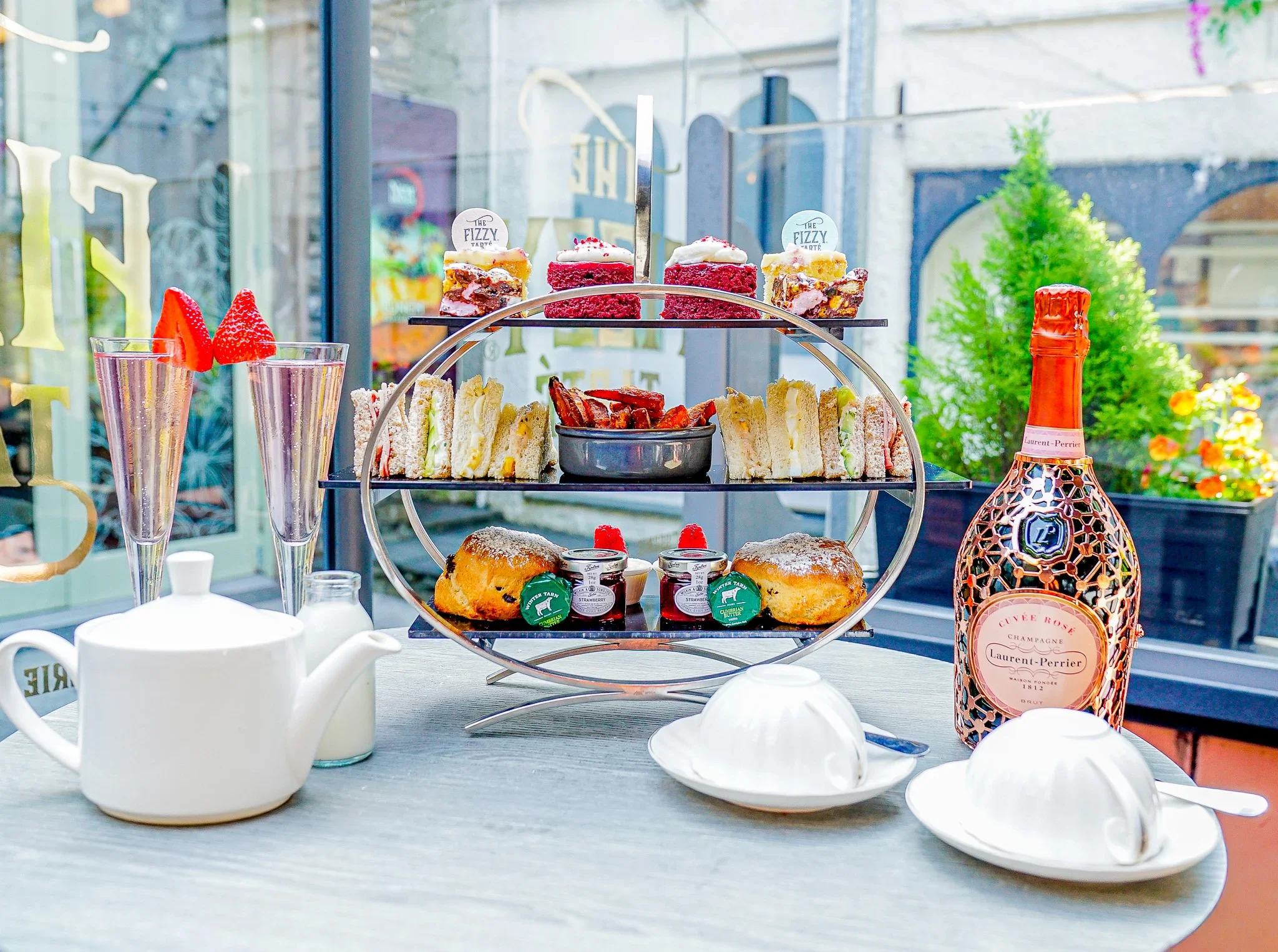 A complete three-tiered Laurent Perrier afternoon tea stand on a round table at The Fizzy Tarté in Bowness-on-Windermere, featuring cakes, sandwiches, sweet potato fries, scones, and tea sets