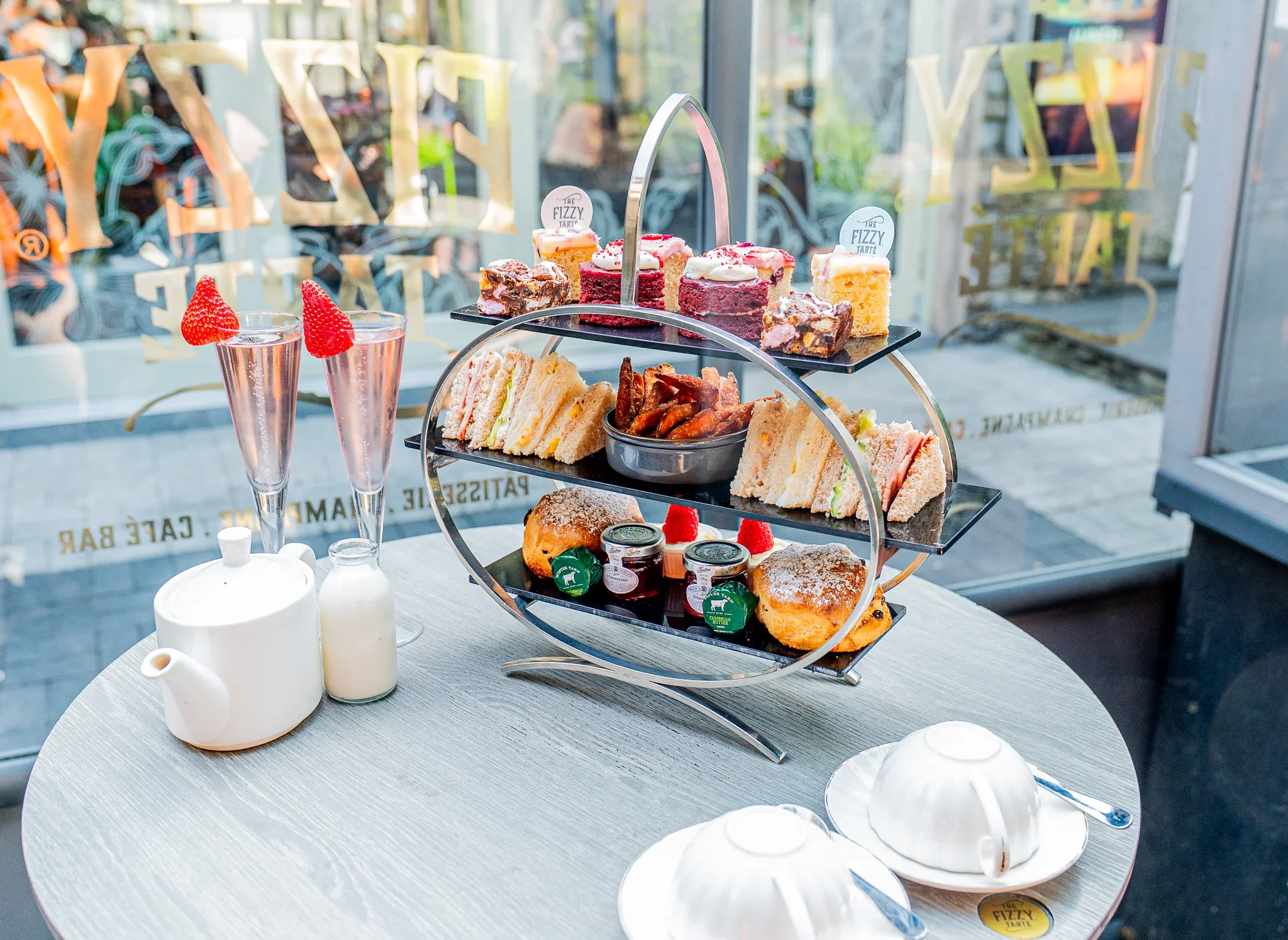 The Ultimate Afternoon Tea experience at The Fizzy Tarté in Bowness-on-Windermere, featuring a three-tiered luxury food stand, tea service, and two tall flutes of pink sparkling Champagne garnished with fresh strawberries.