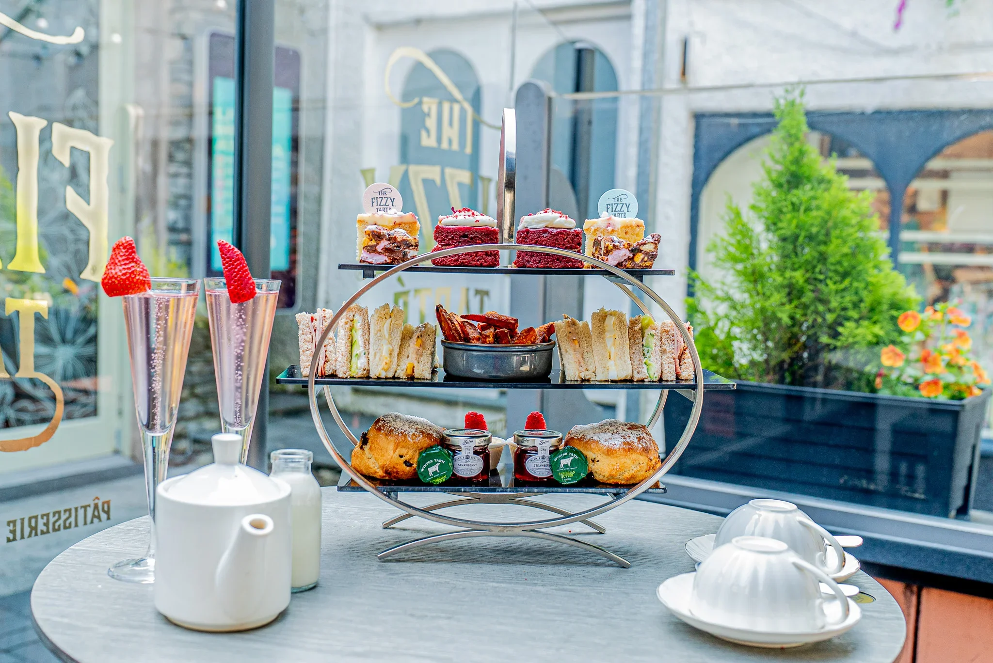 The Ultimate Afternoon Tea experience at The Fizzy Tarté in Bowness-on-Windermere, featuring a three-tiered luxury food stand, tea service, and two tall flutes of pink sparkling Champagne garnished with fresh strawberries.