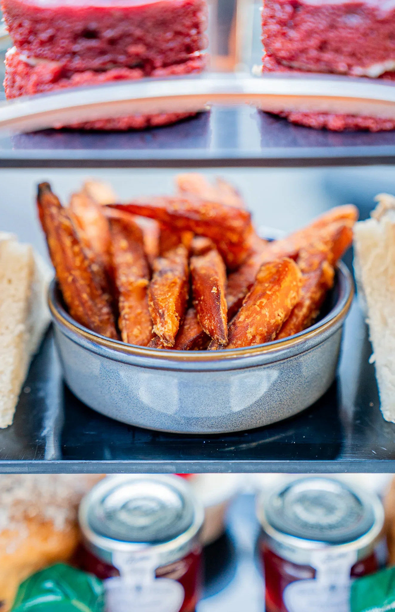 Close-up of an afternoon tea stand tier at The Fizzy Tarté in Bowness, showing a ceramic bowl filled with golden, crispy sweet potato fries nestled between fresh finger sandwiches, with red velvet cakes visible on the tier above.