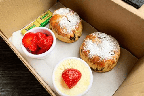 Box containing two muffins dusted with powdered sugar, a small bowl of strawberries, a small bowl of pudding with a strawberry on top, and two small packets of butter.