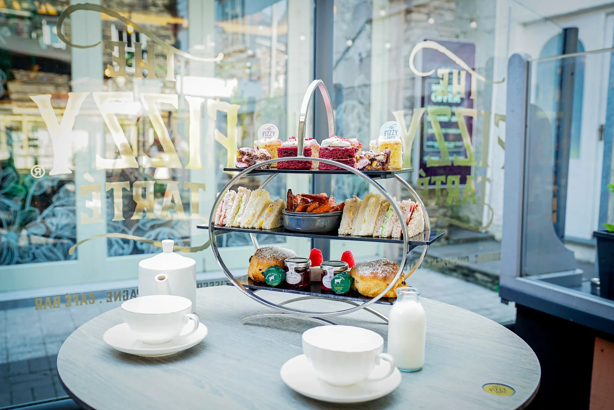 A classic three-tiered afternoon tea stand on a round outdoor table at The Fizzy Tarté in the Lake District, featuring cakes, finger sandwiches, sweet potato fries, scones, and a white porcelain tea set.