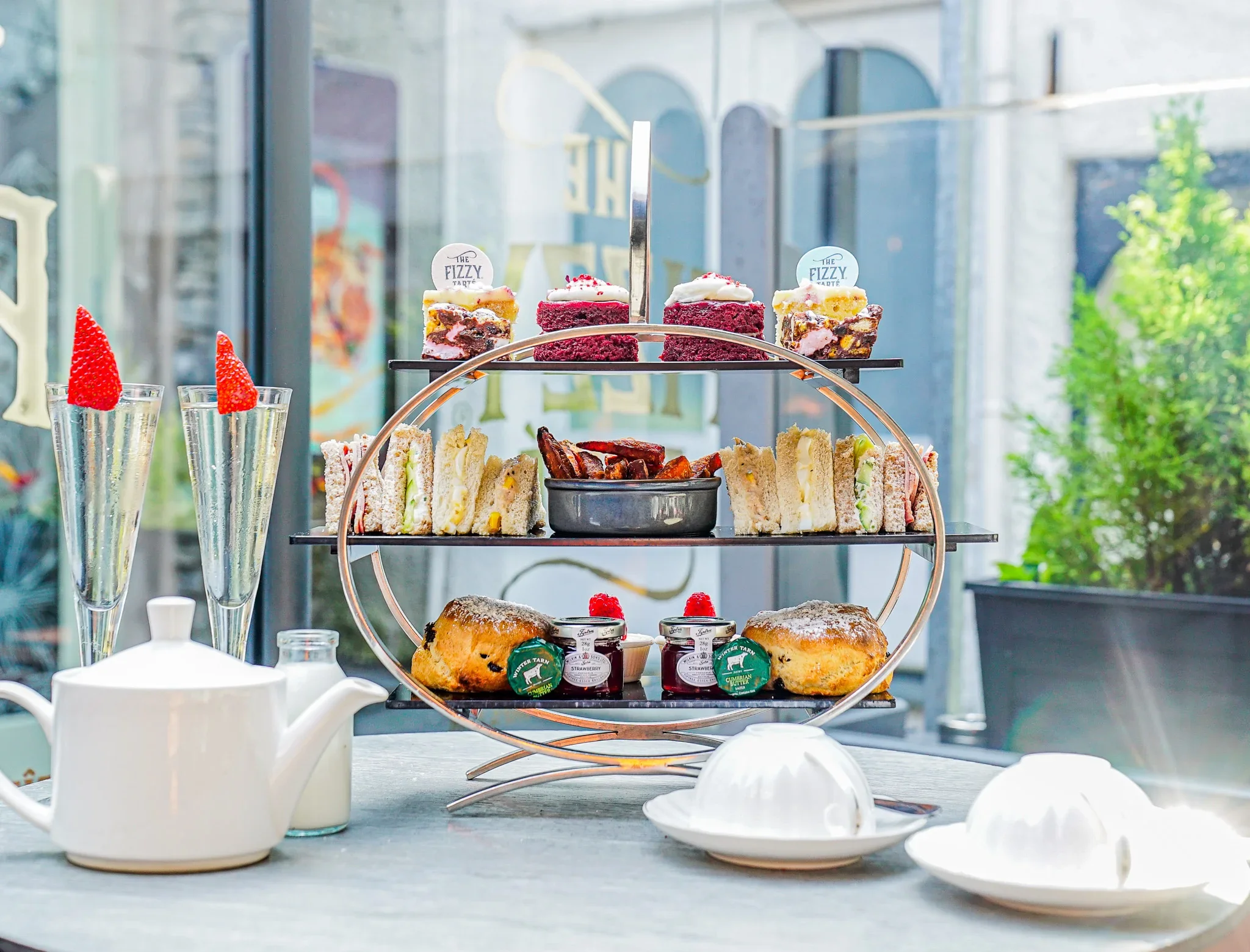 The Ultimate Afternoon Tea experience at The Fizzy Tarté in Lake District , featuring a three-tiered luxury food stand, tea service, and two tall flutes of white sparkling Champagne garnished with fresh strawberries.
