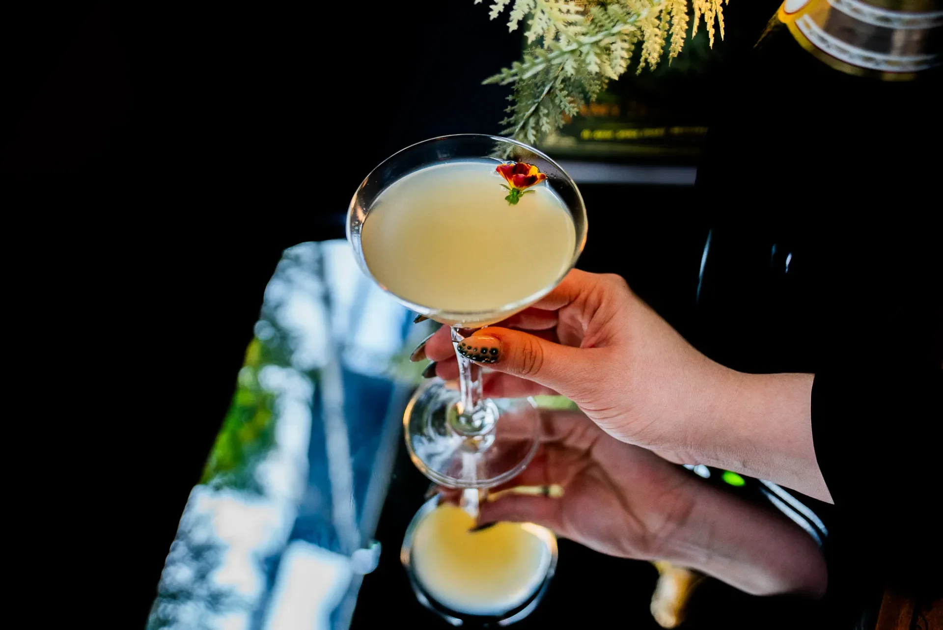 A hand holding a cocktail glass filled with a pale yellow drink, garnished with a small edible flower.