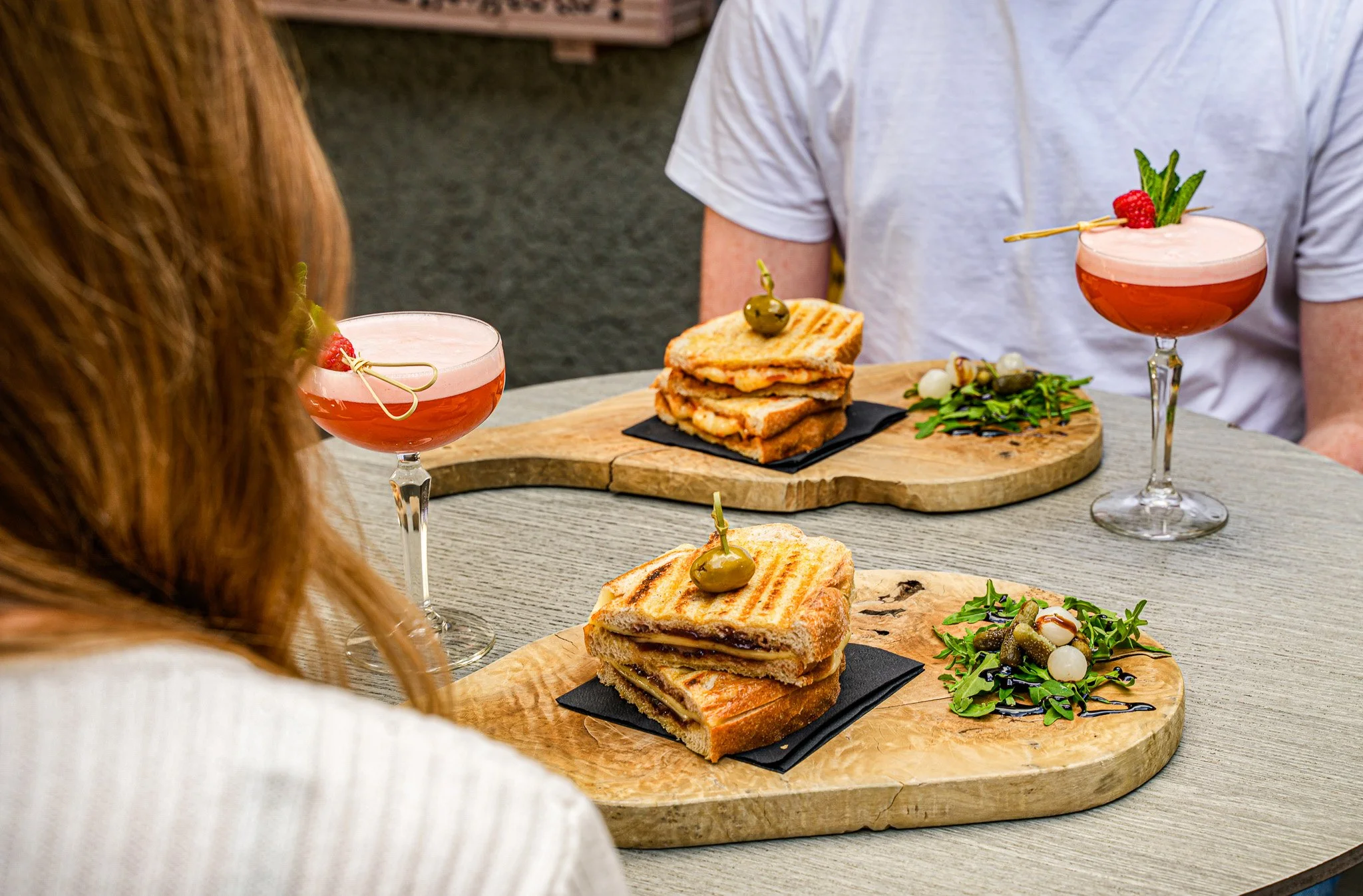 Two people sitting at a table with wood serving boards holding grilled sandwiches, green salad with balsamic drizzle, and pink cocktails garnished with raspberries, on a light wood table.