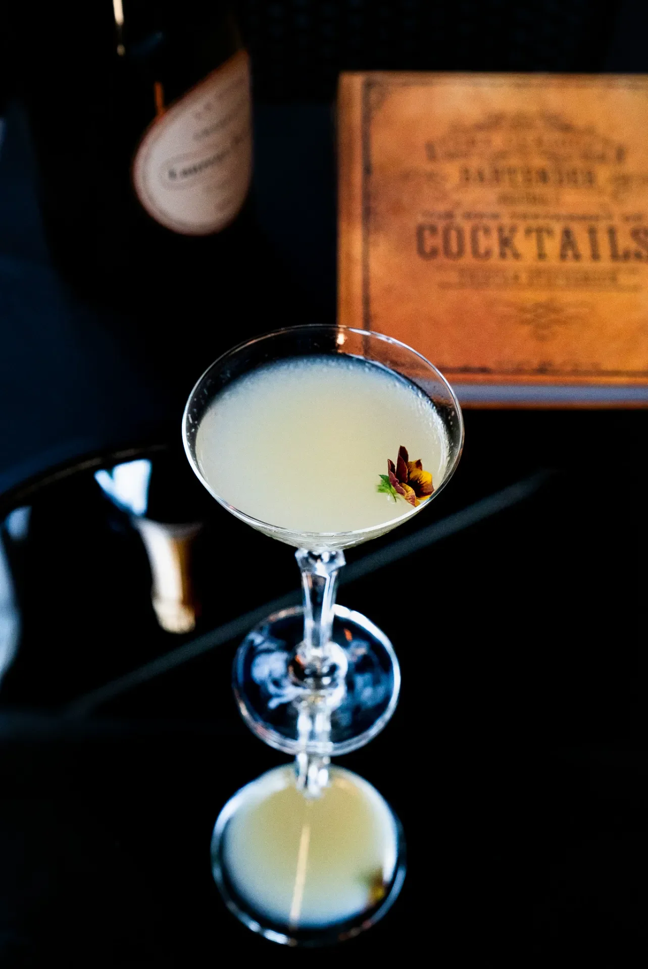A cocktail glass filled with a pale yellow drink, garnished with a small edible flower. In the background, there are two bottles and a wooden menu with the word 'COCKTAILS' on it.