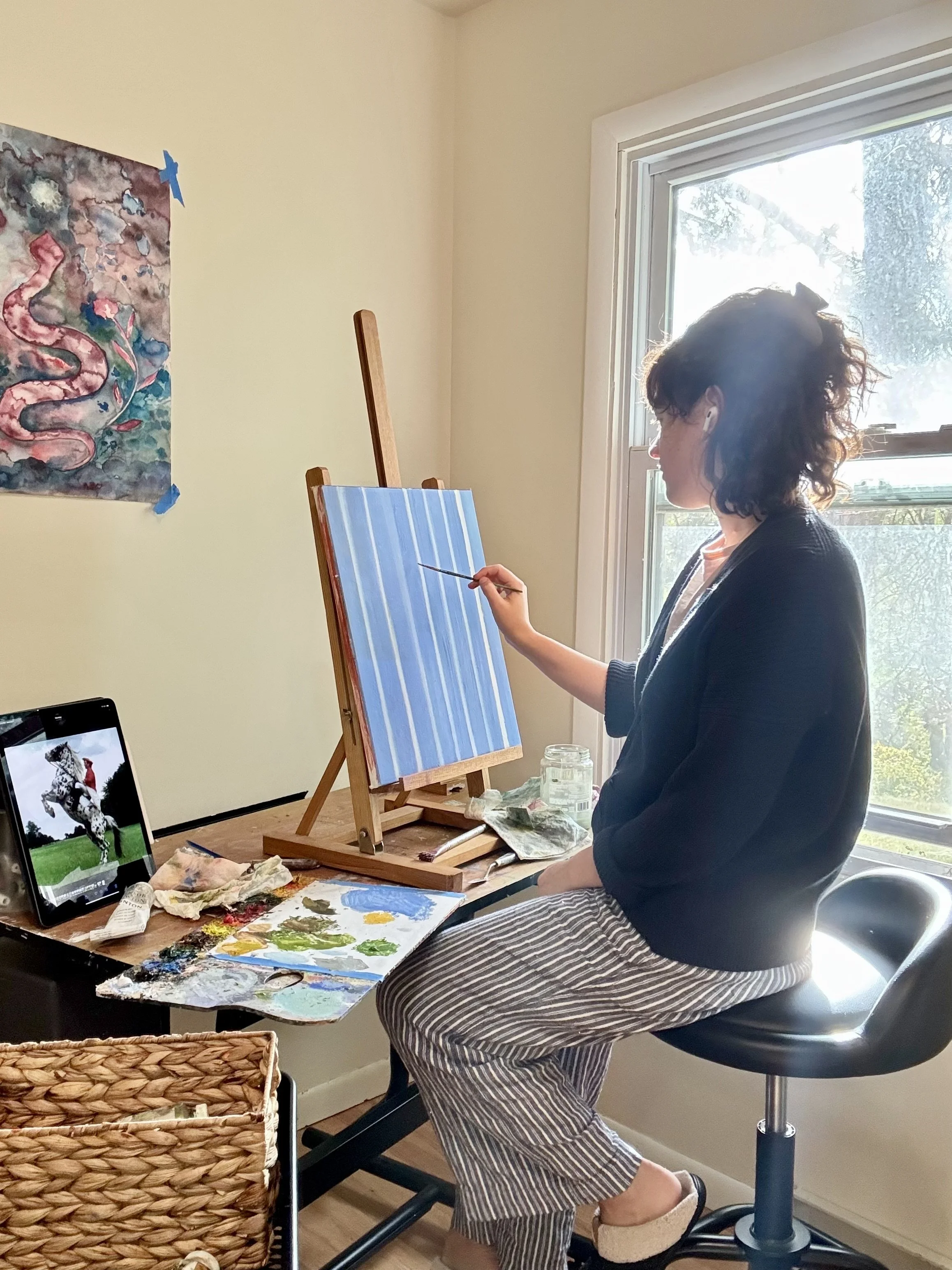A woman painting a blue and white striped canvas on an easel in a bright room with large window, sitting on a black stool, with art supplies and a tablet displaying a horse on the desk.