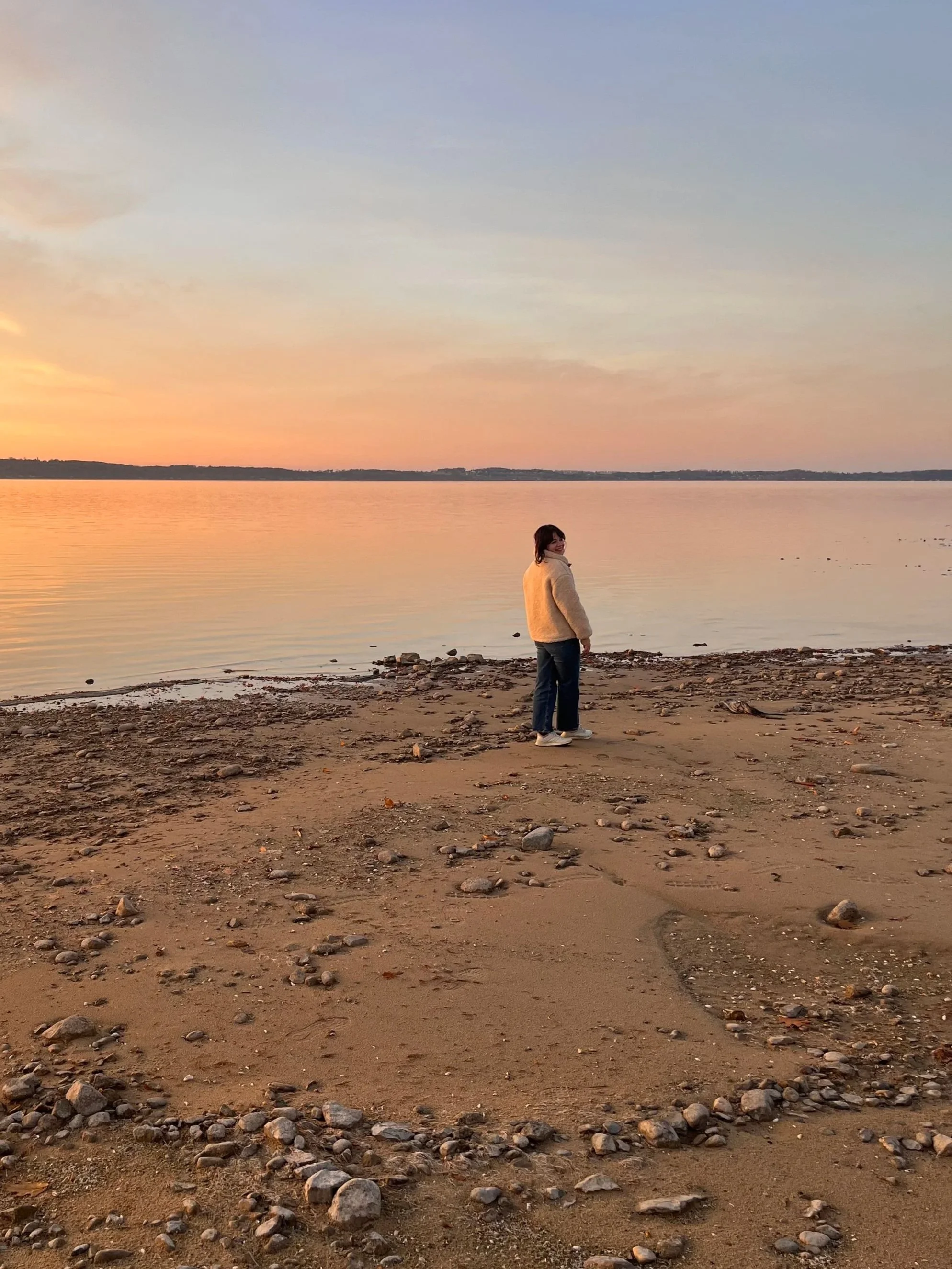 Person standing on a sandy beach near a body of water during sunset, with a partly cloudy sky.