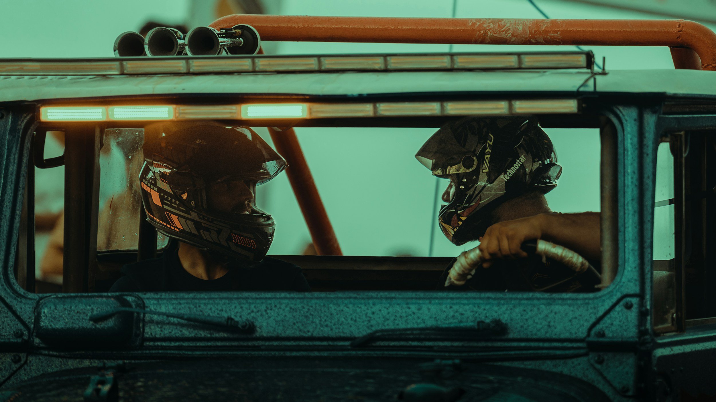 Two race car drivers wearing helmets sit inside a race car, facing each other, possibly having a conversation before a race.