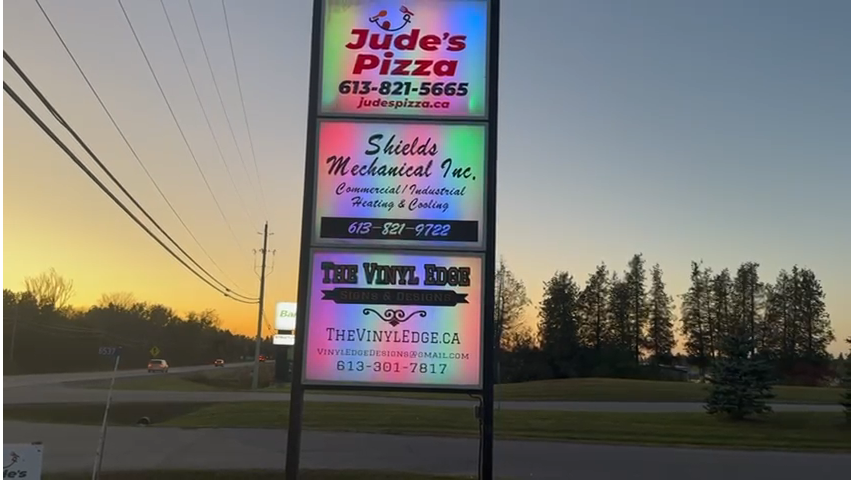 Outdoor sign featuring businesses: Jude's Pizza, Shields Mechanical Inc., and The Vinyl Edge, set against a sunset background.