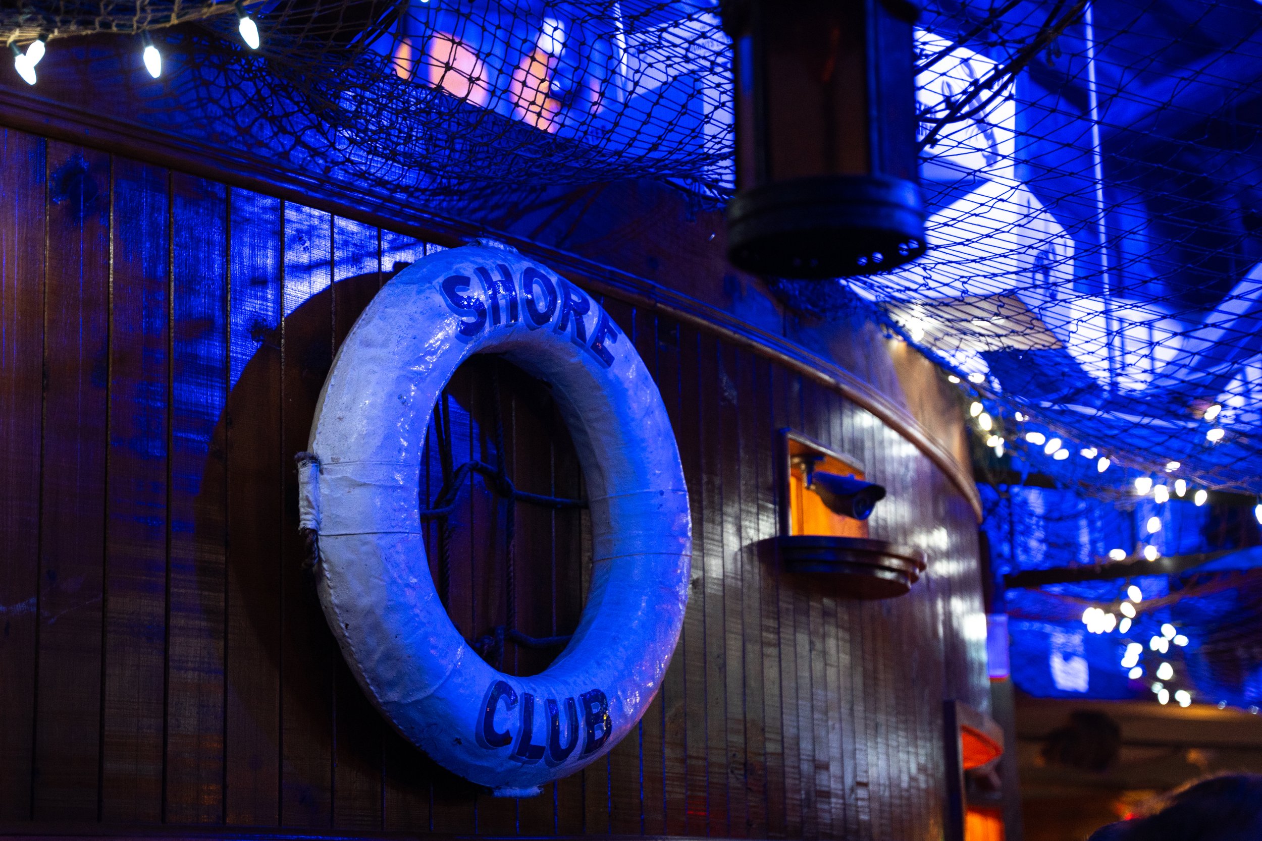 Interior of a bar with nautical decor, featuring a lifebuoy with "Shore Club" written on it, netting, and decorative lights.