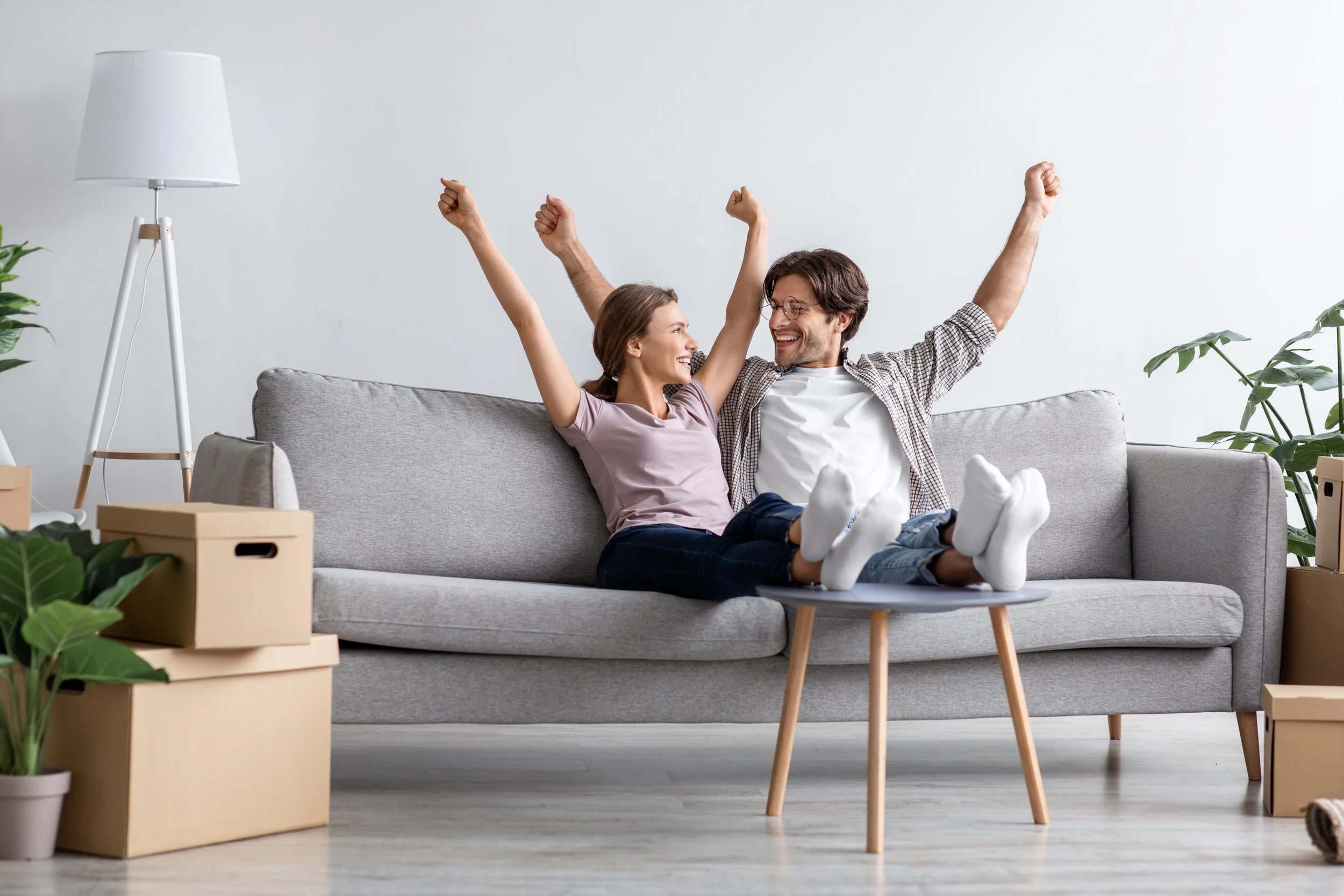 A happy couple sitting on a light gray sofa with small wooden legs in a bright living room, celebrating with raised fists and smiling. There are green plants and cardboard boxes around them, suggesting they are moving or unpacking.