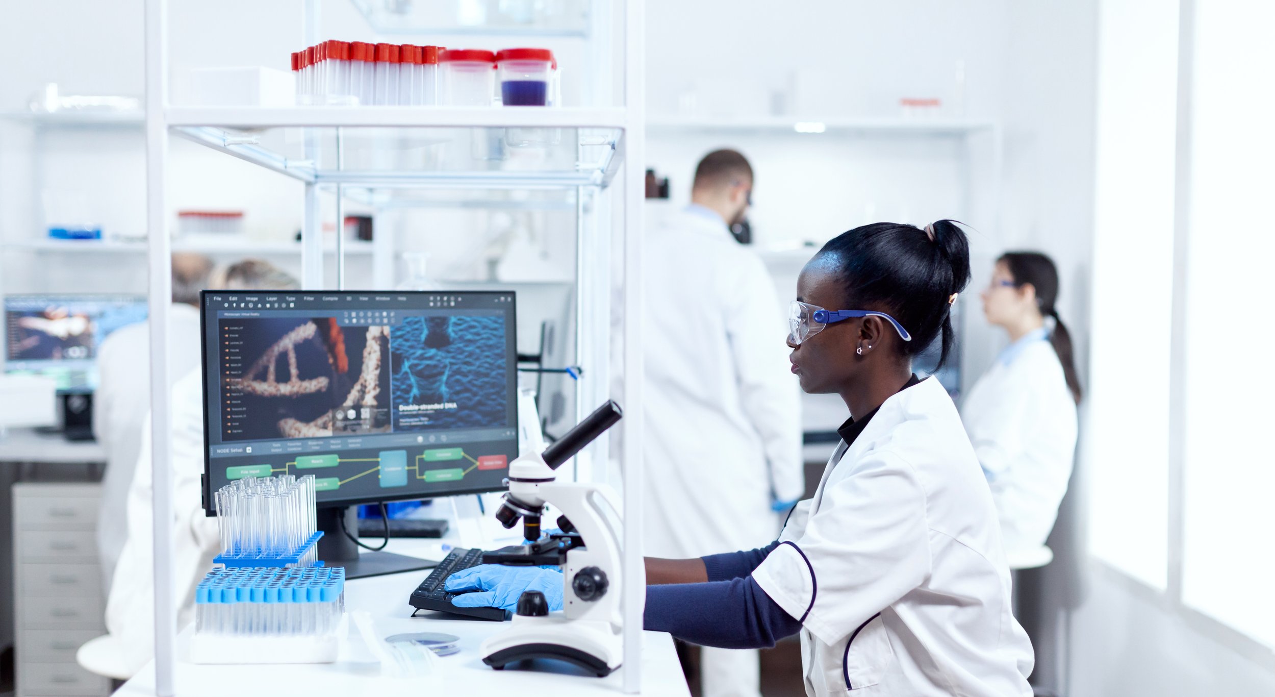 Female scientist looking at her computer screen showing DNA and laboratory results in a modern laboratory with other researchers working in the background.