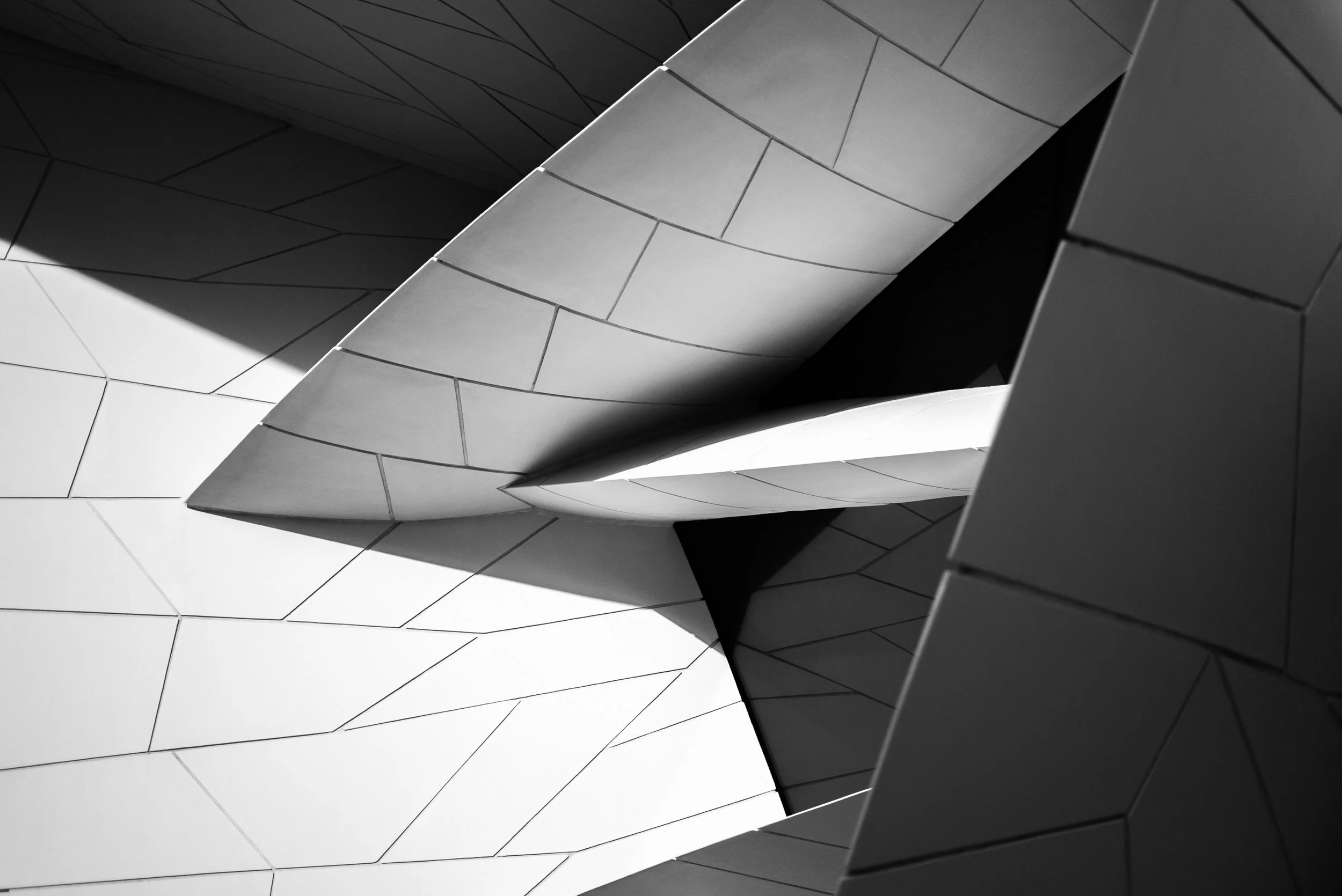 Close-up of a modern building's architecture with angular, geometric, metallic surfaces and contrasting shadows in black and white.