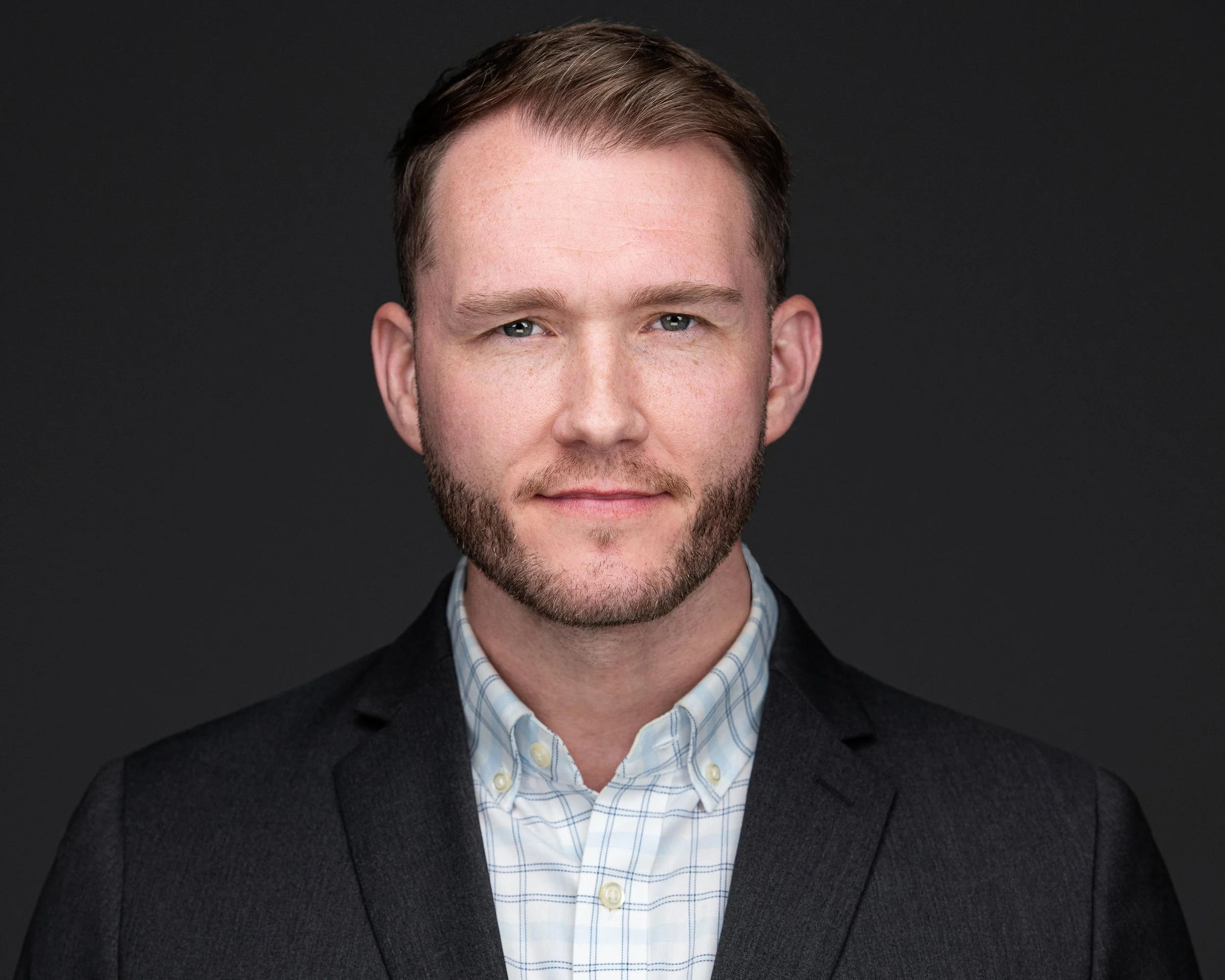 Close-up of a man with short brown hair, light skin, and blue eyes, wearing a checkered shirt and a dark blazer, against a black background.