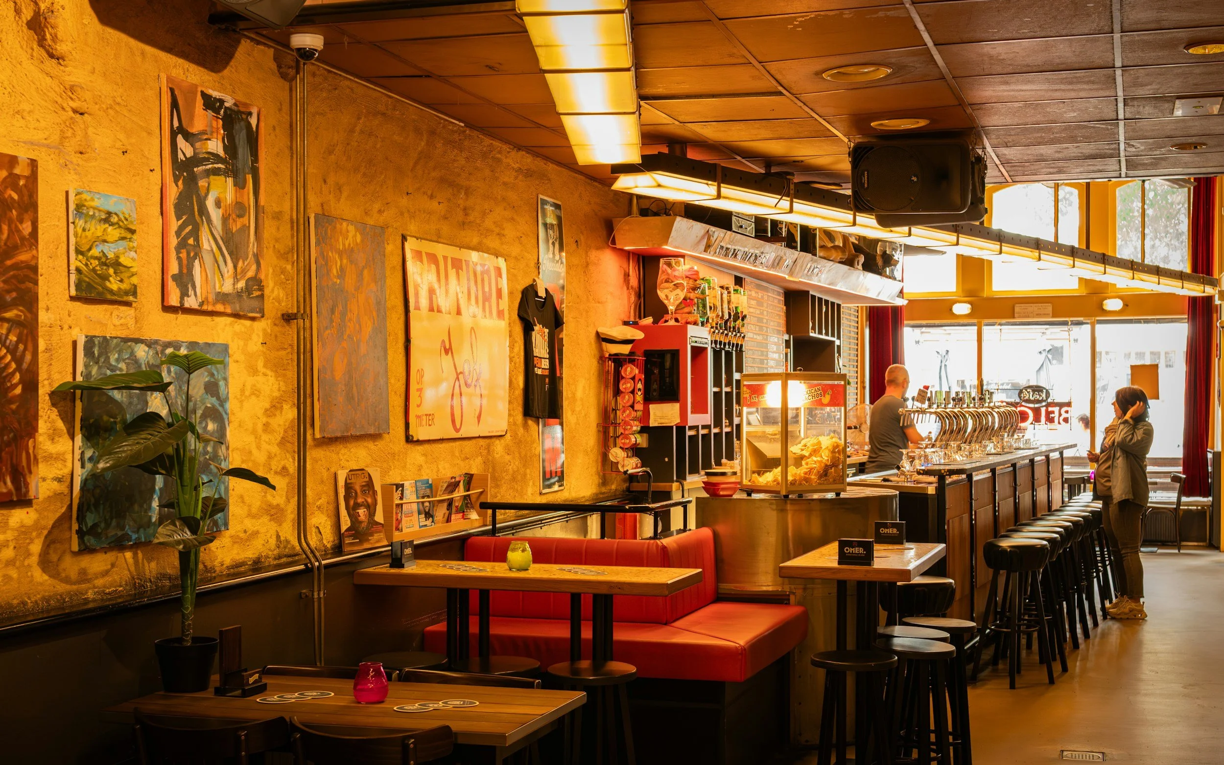 Interior of a restaurant with artwork on a yellow brick wall, red booth seating, and bar stools along a counter, with two people near the counter and large windows in the background.