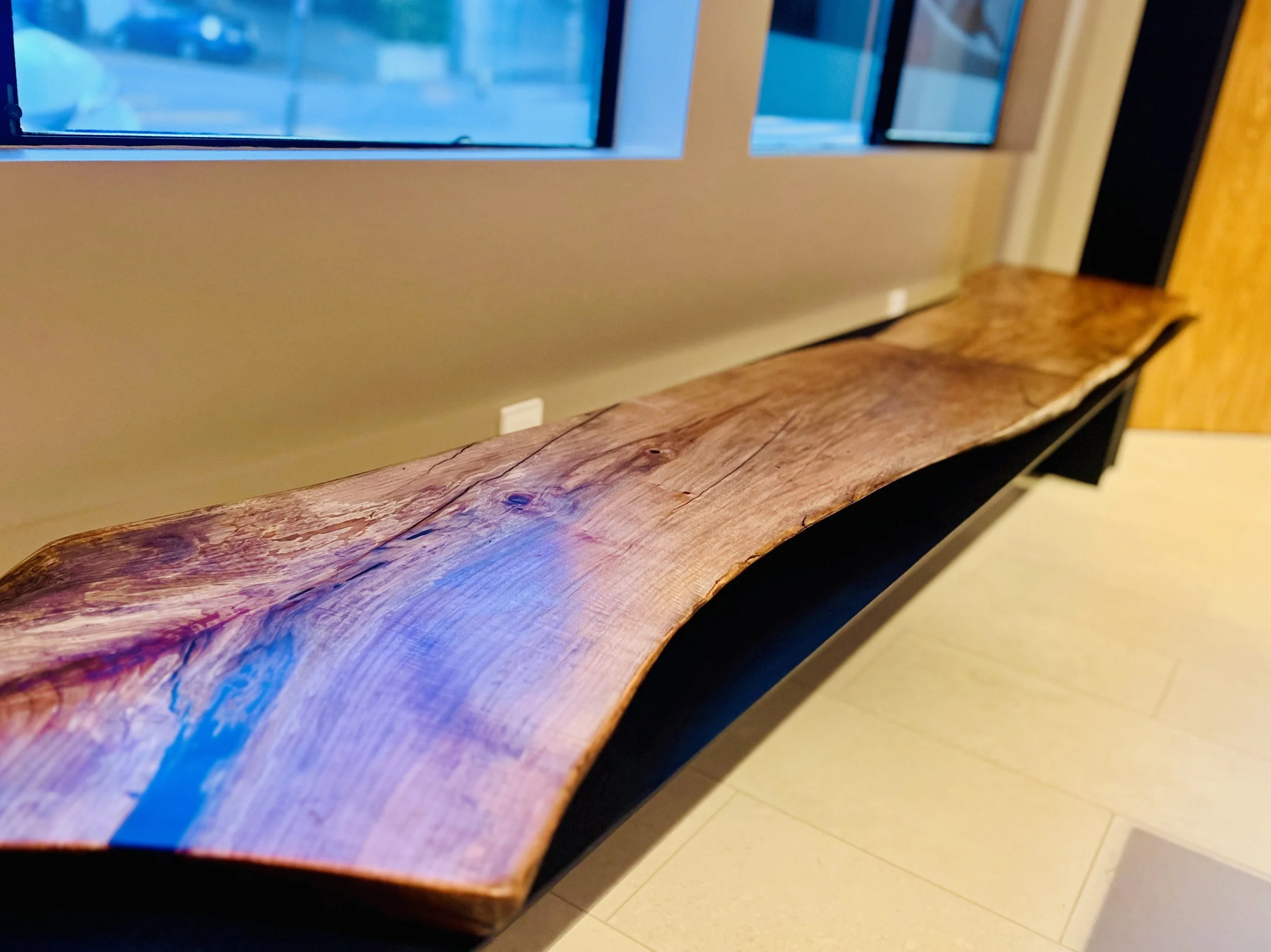 A long, curved wooden bench with a natural, unpolished finish positioned below a large window in a room with beige walls and light-colored tiled flooring.