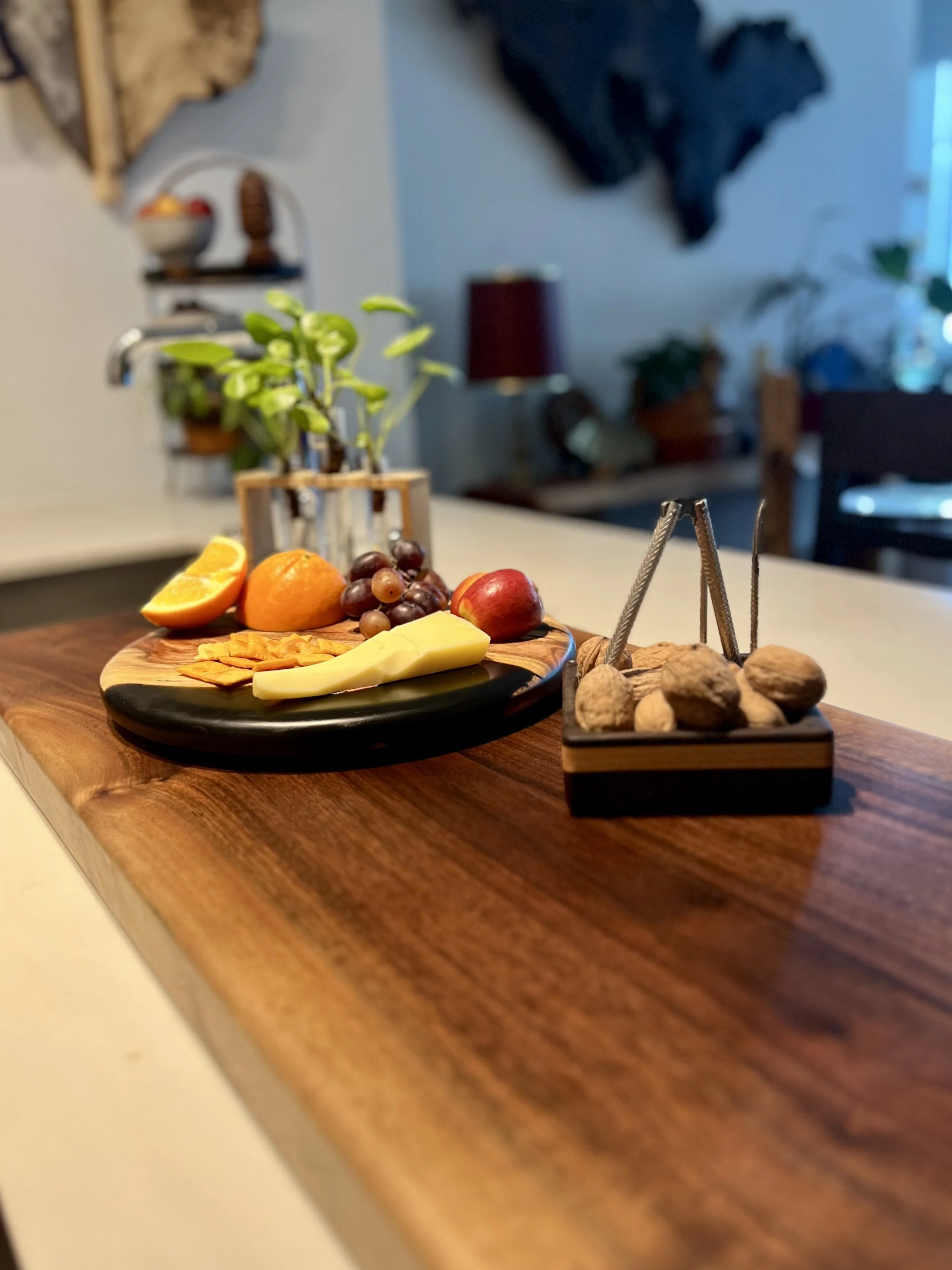 A wooden countertop with a black plate holding sliced orange, an orange, grapes, apple, and cheese. Next to the plate is a small box of nuts with tongs, and a potted plant in the background.