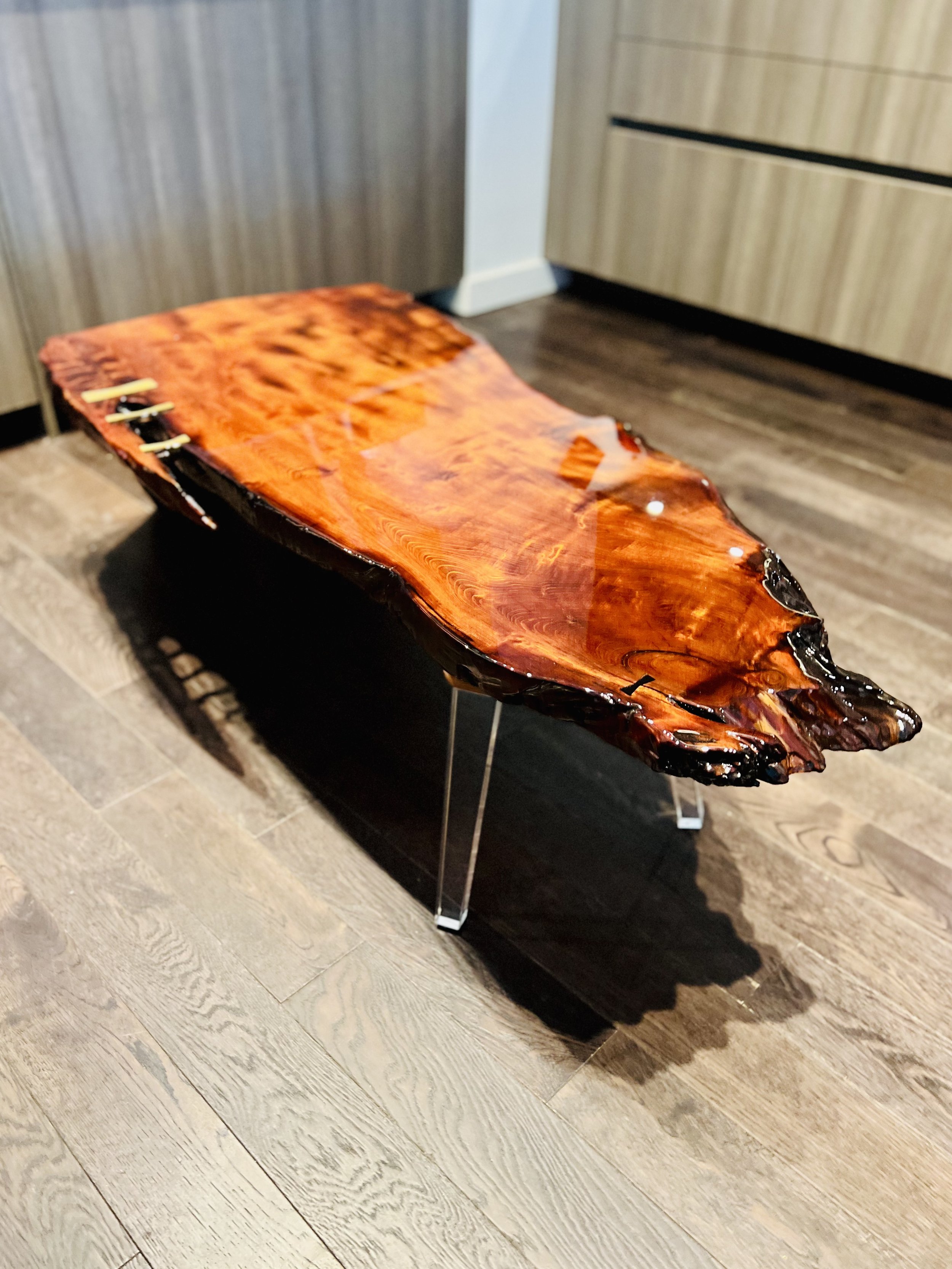 A polished wooden surfboard-shaped table with a glossy finish, supported by metal hairpin legs, placed on a hardwood floor.