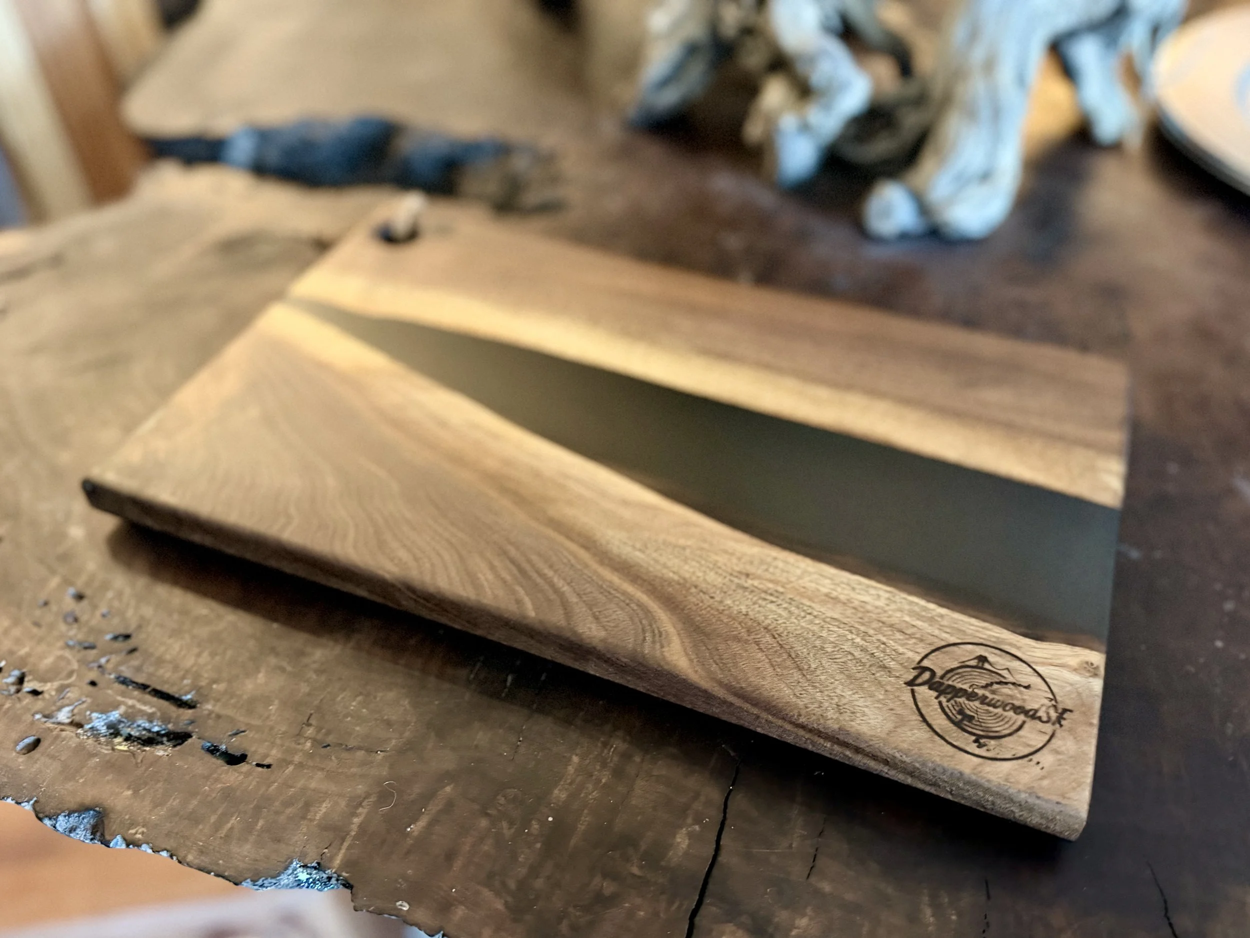 A wooden serving tray with a resin center, placed on a rustic wooden table. The tray has a logo in the corner that reads "Dapperwood" with a mountain and tree design.
