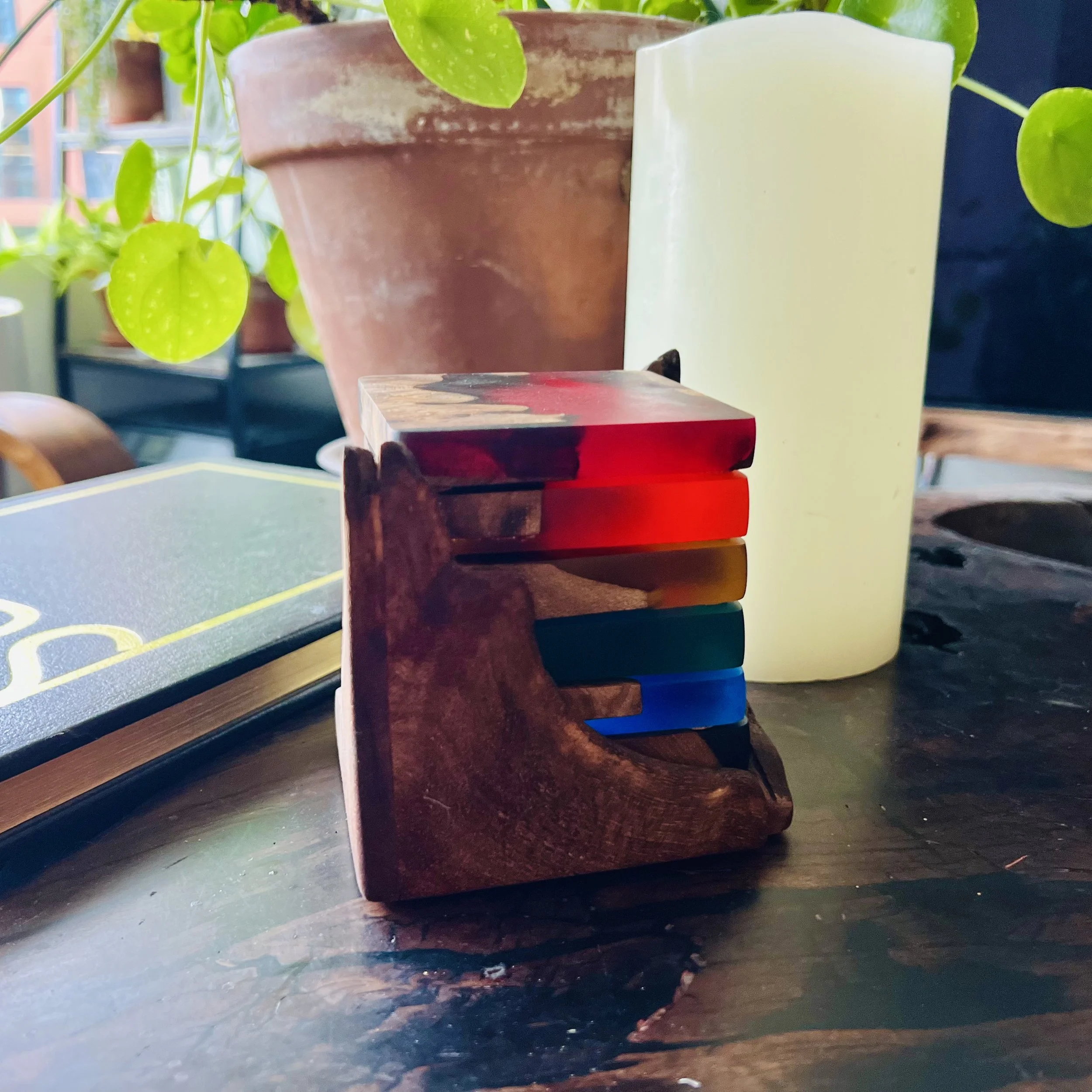 A stack of multicolored oasters on a wooden stand, next to a large potted plant, a candle, and a closed book on a dark wooden table.