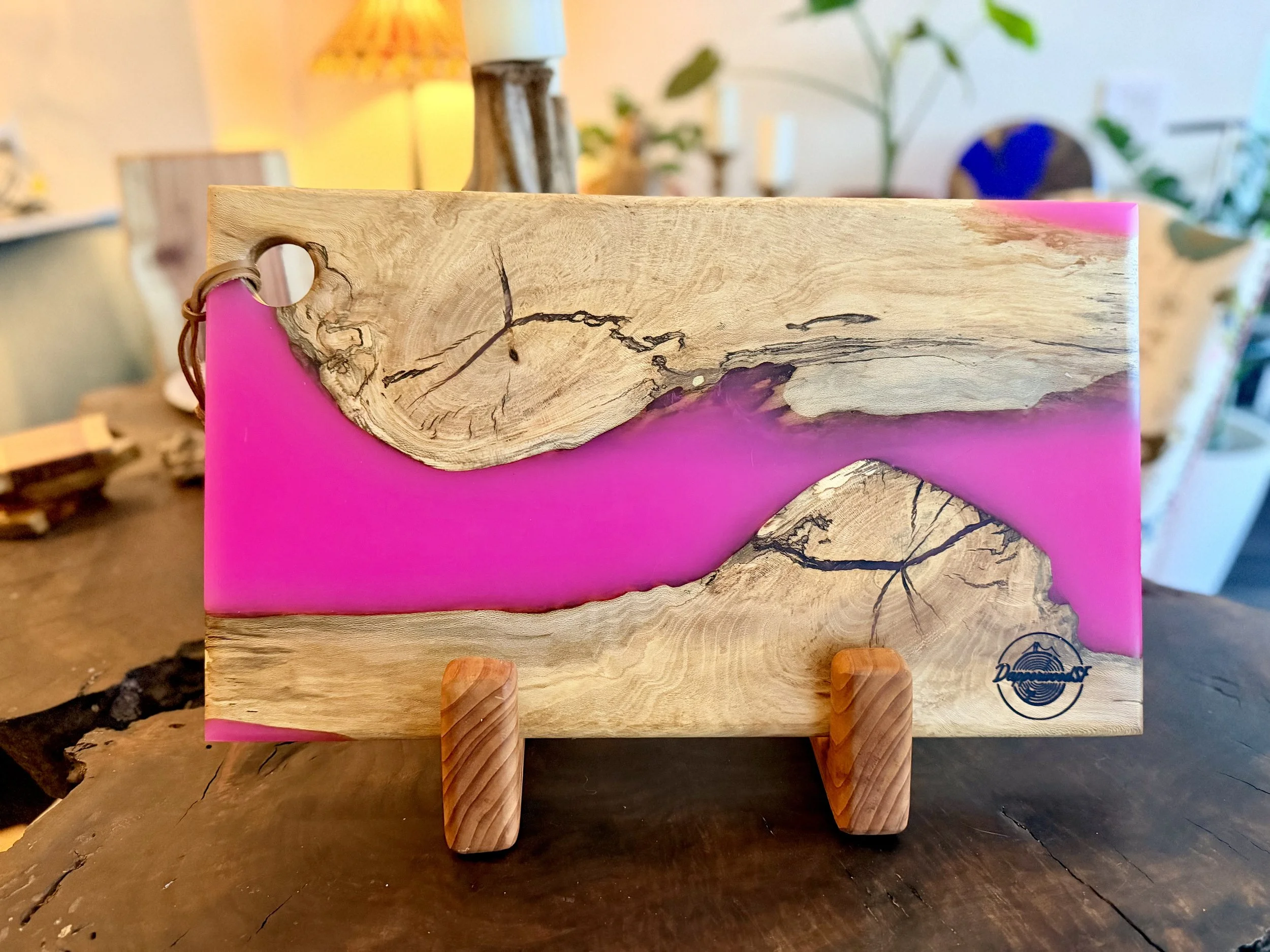 A decorative wooden plaque with pink resin accents and black cracks, displayed on wooden stands on a dark wooden table.