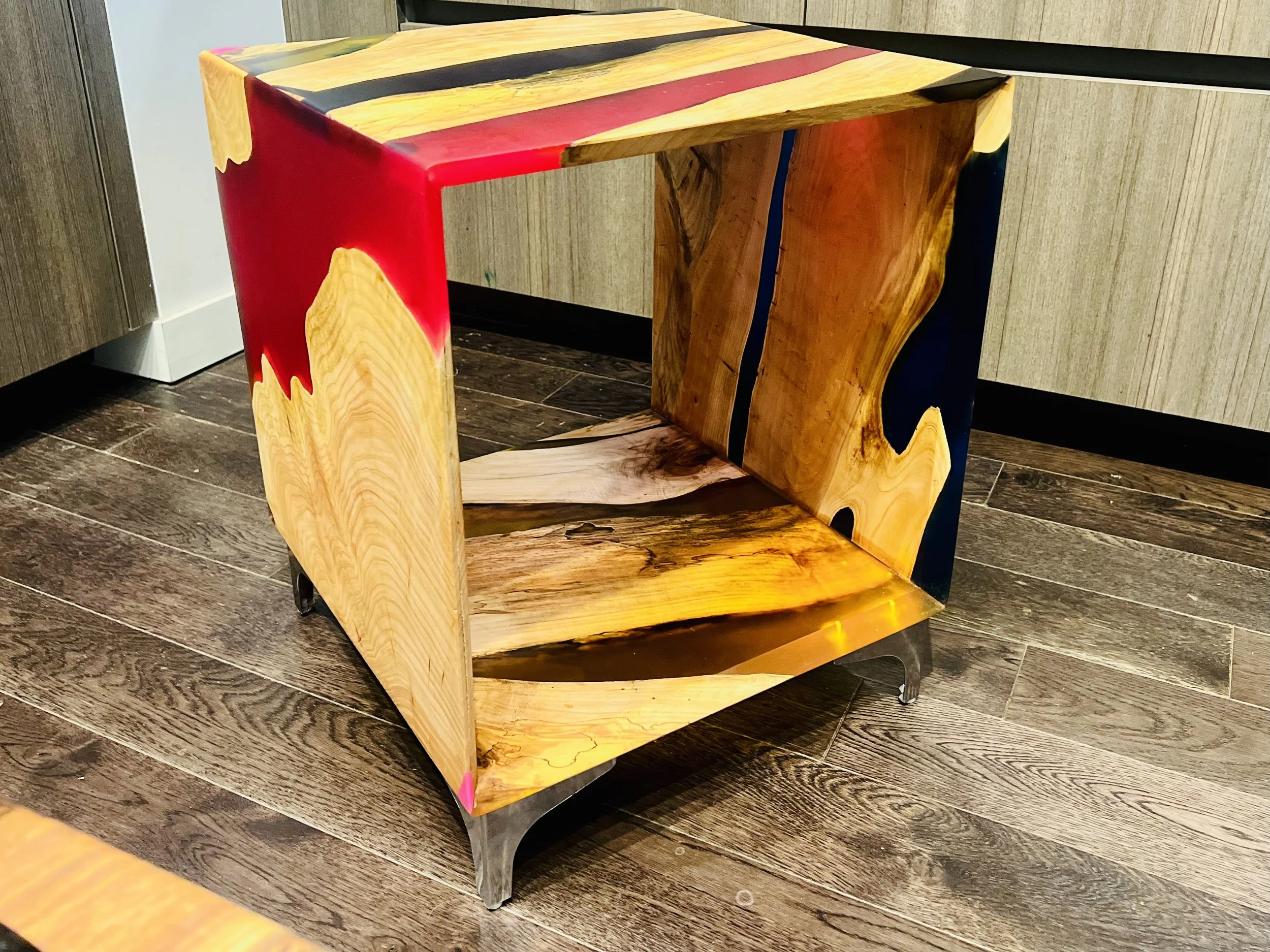 A wooden cube-shaped side table with a unique design, featuring natural wood grain with colored paint accents in red, blue, and pink, on a brown wood floor.