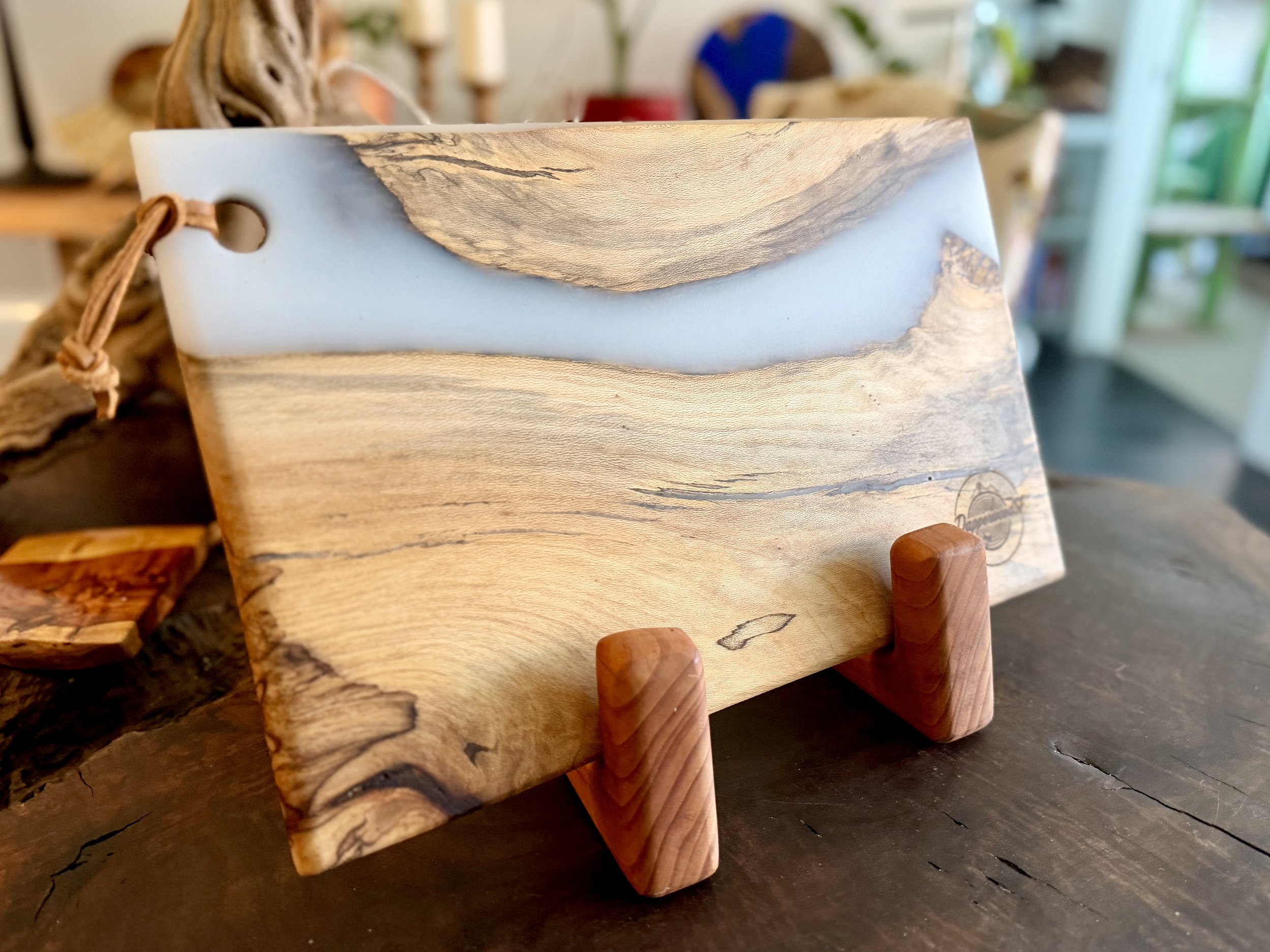 A wooden cutting board with a light-colored resin inlay, displayed on a wooden stand in a craft shop.