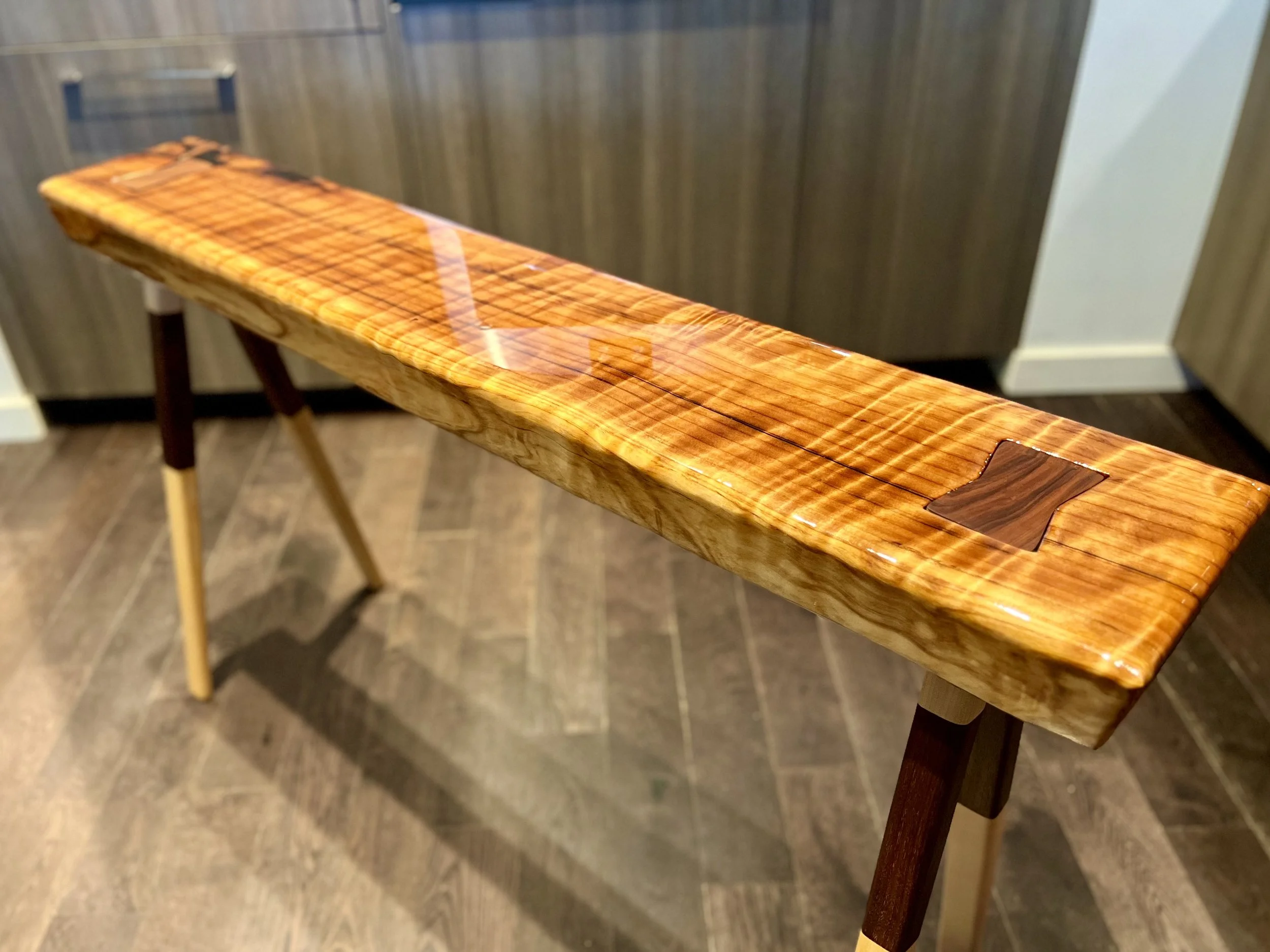 Wooden bench with a glossy finish and a rectangular inlay in the center, placed on a hardwood floor in front of wooden cabinets.