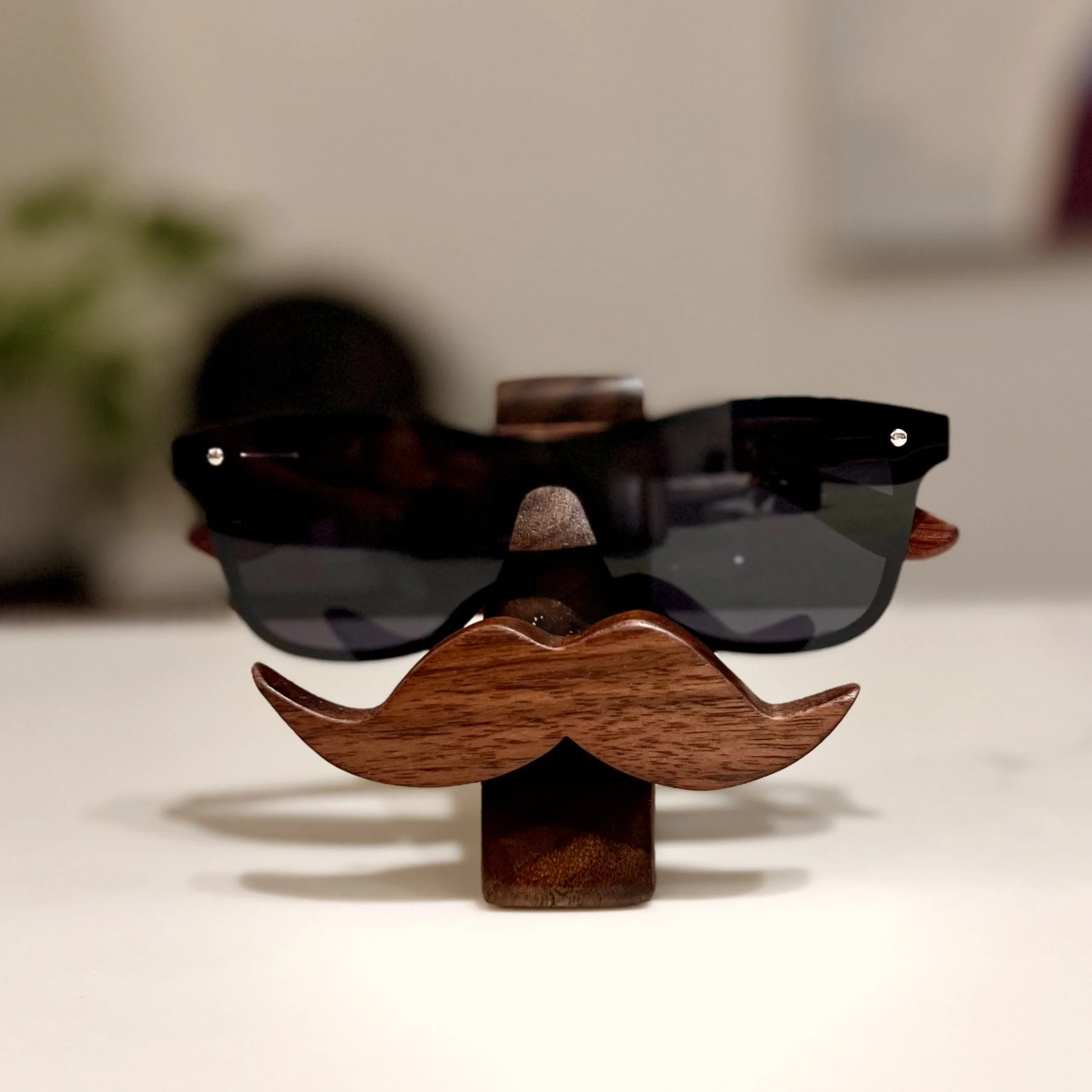 A wooden mustache stand with black sunglasses placed on top, on a white surface with a blurred background.