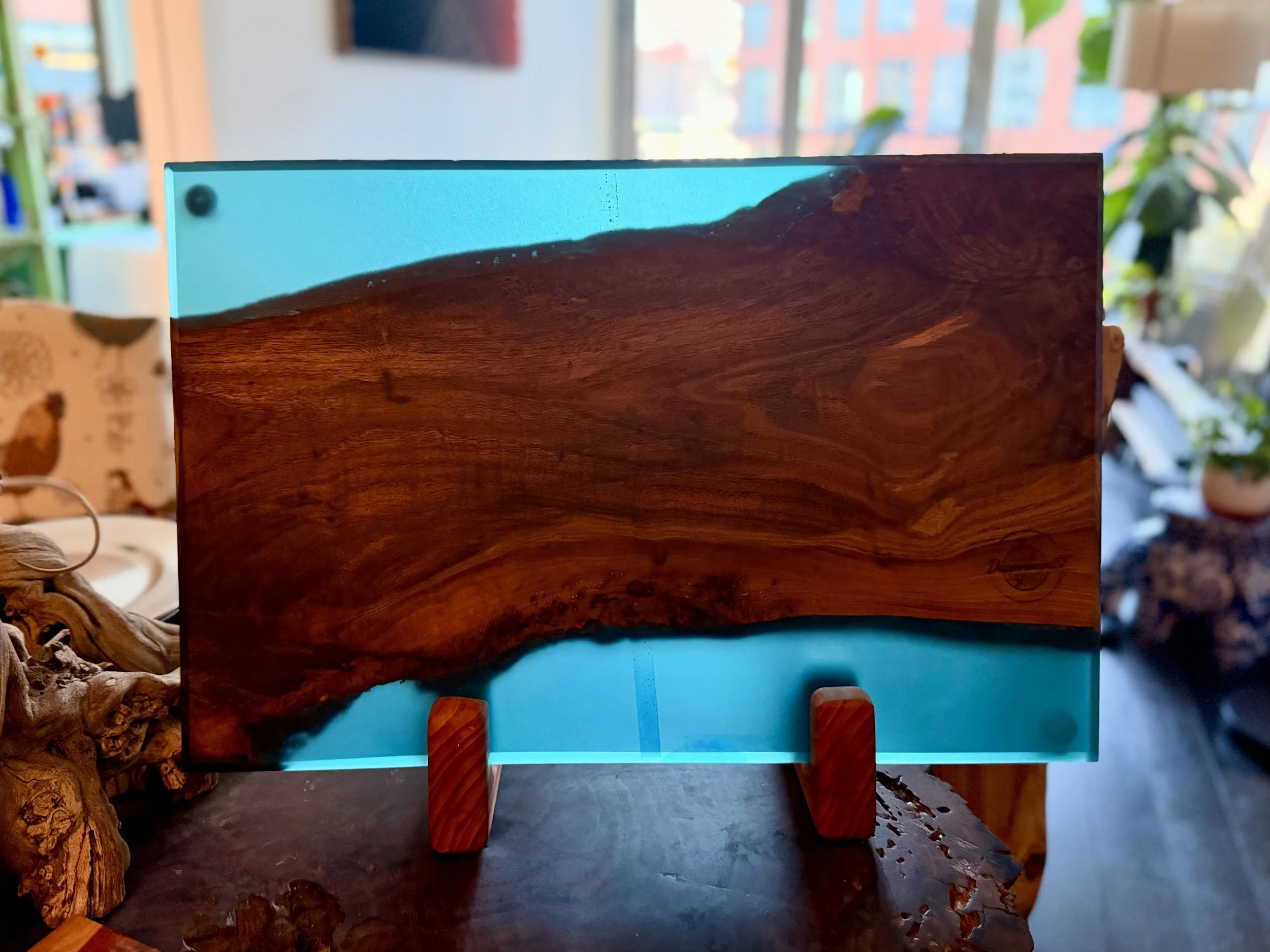 A decorative art piece with a large piece of wood mounted on a light blue glass base, displayed on a wooden surface inside a room with large windows, potted plants, and various items in the background.