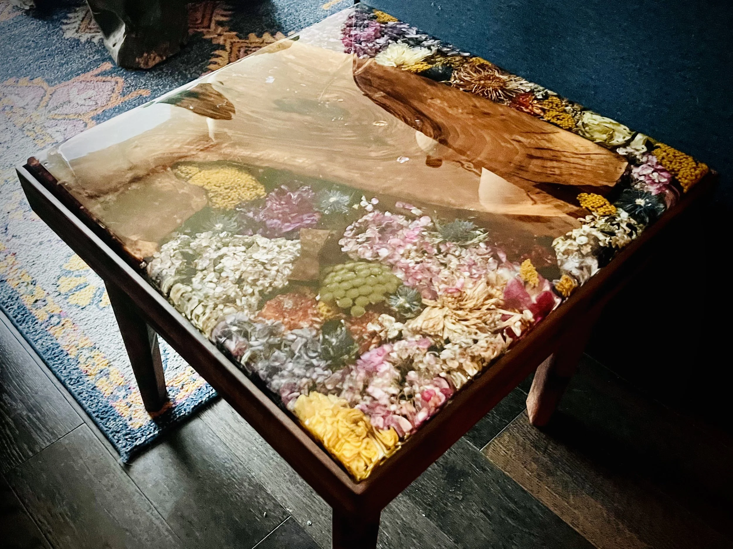 A wooden table with a glass top displaying a variety of dried flowers and plants arranged underneath.
