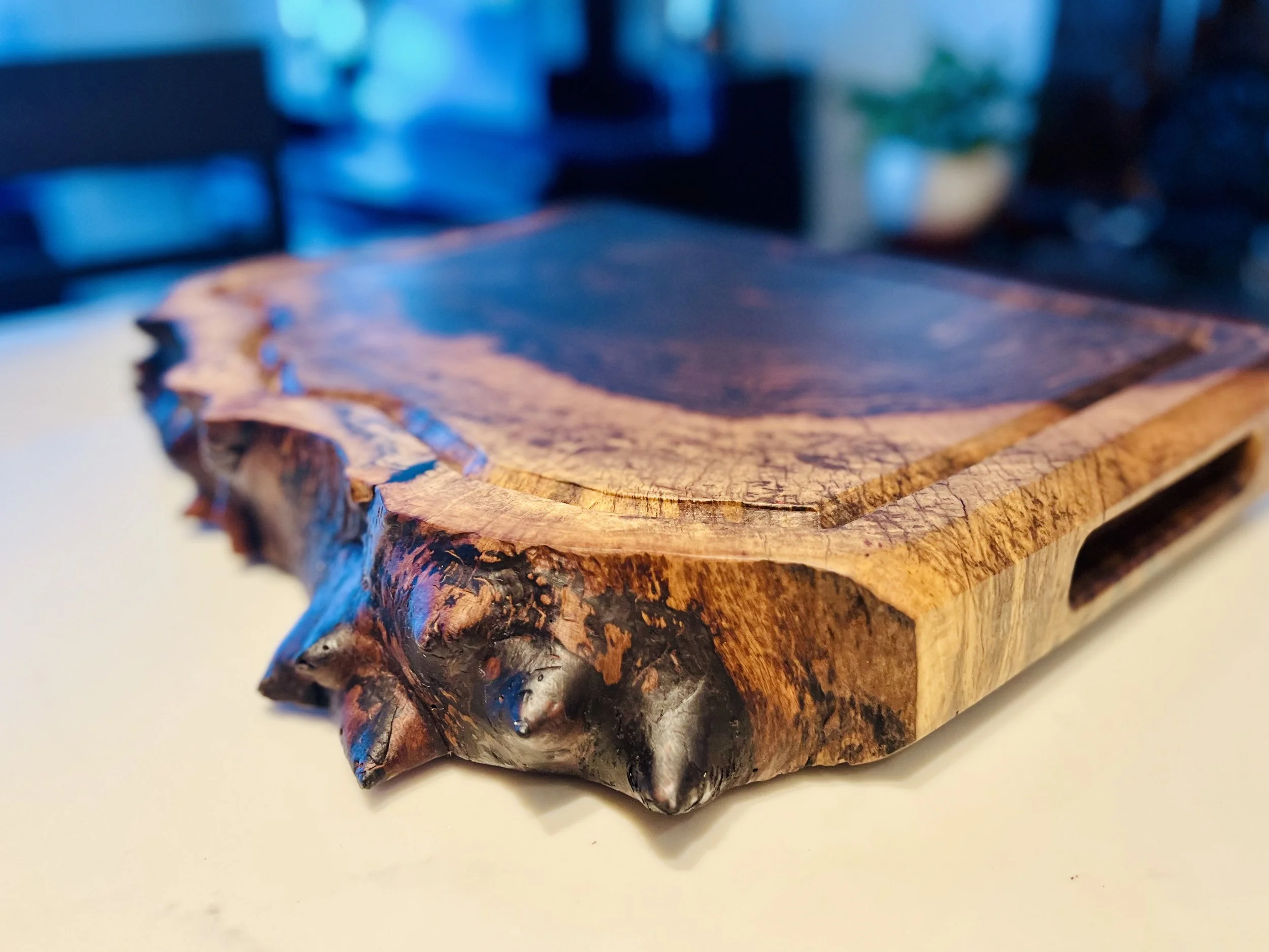 Close-up of a wooden cutting board with a natural, uneven edge, placed on a white surface. The background is blurred with blue and dark tones.
