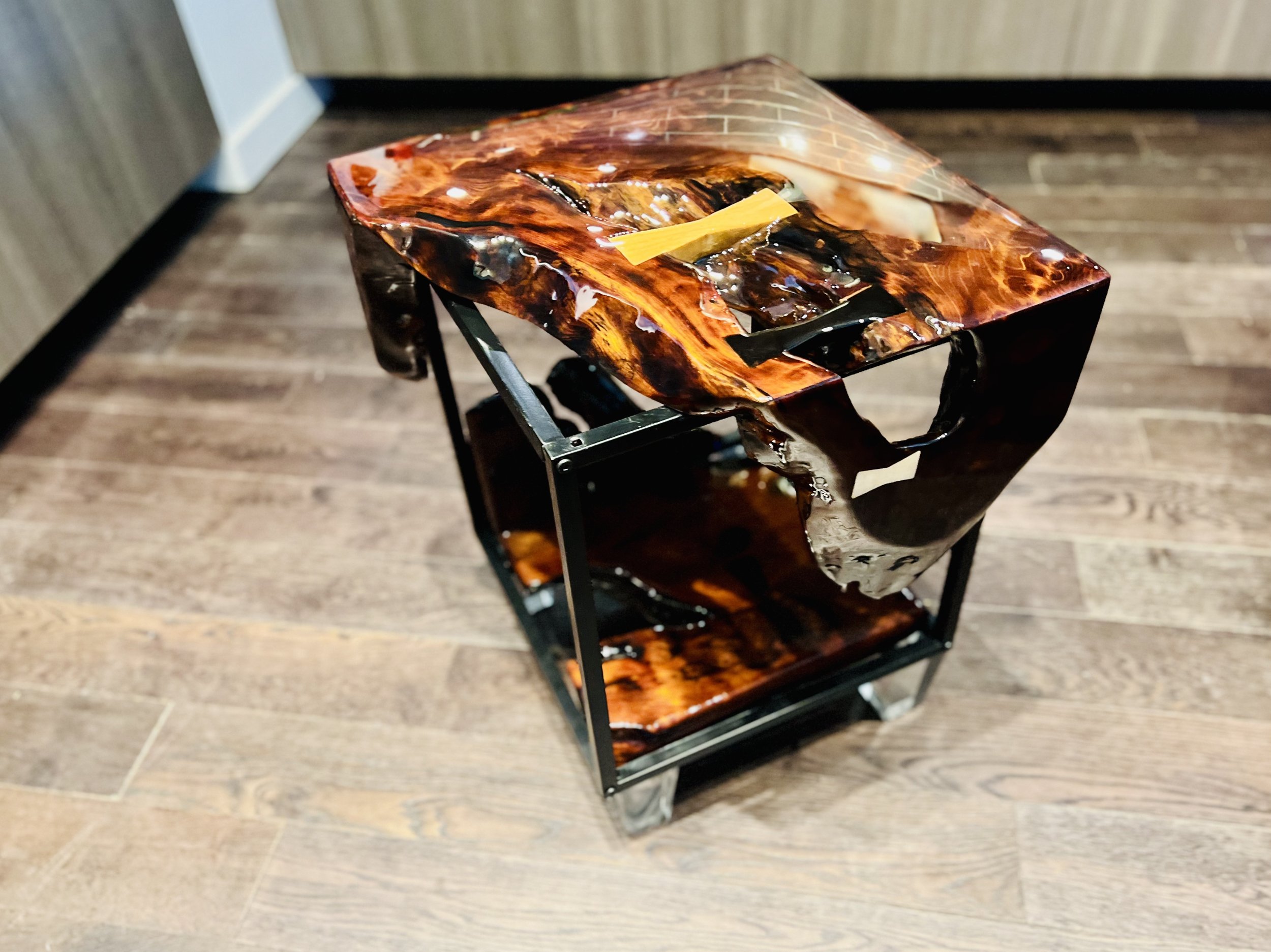 A modern portable fireplace with a glossy, marbled finish in shades of brown and black, on a black metal frame, placed on a wooden floor in a room.