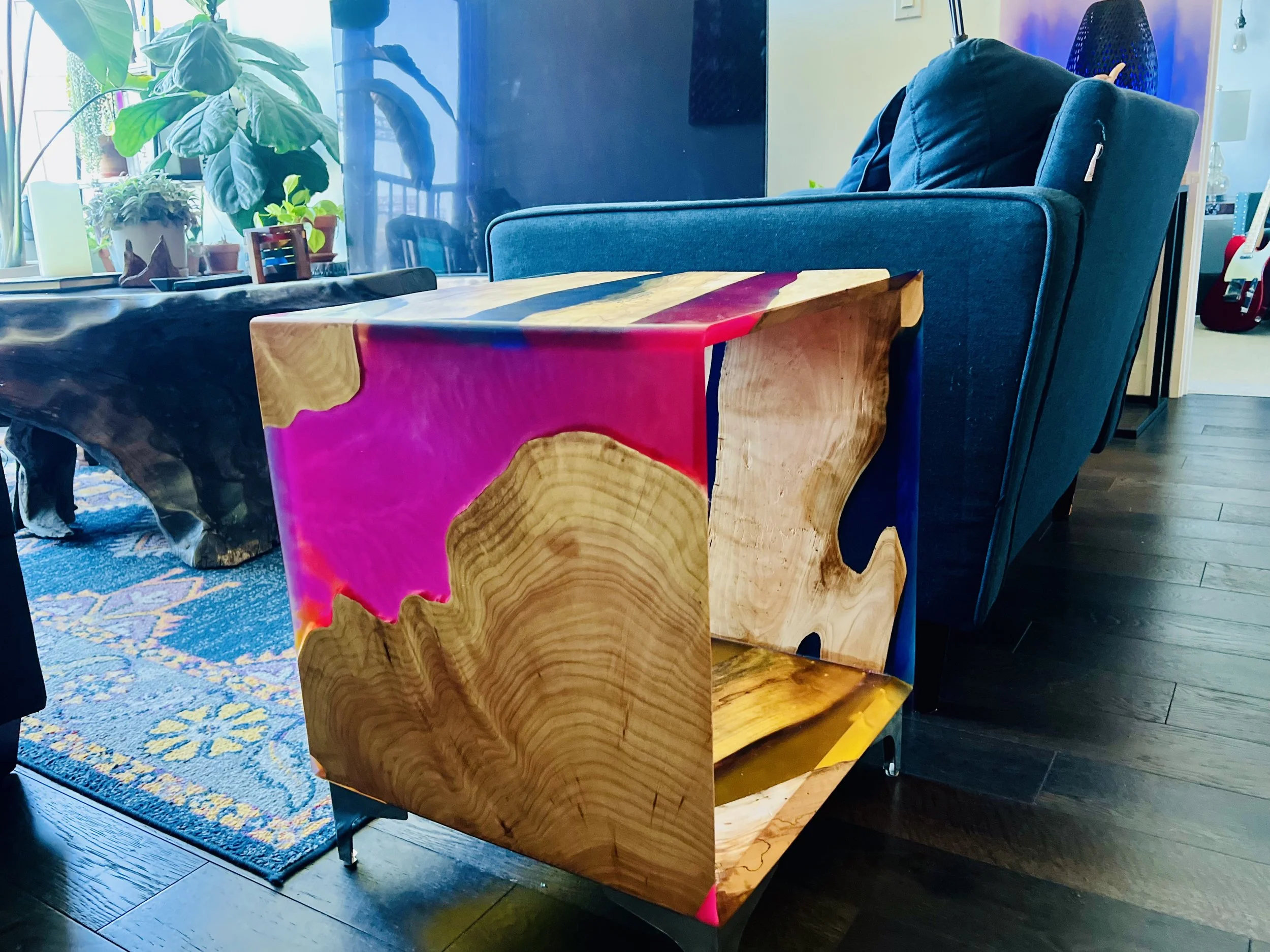 A side table made of wood with pink and blue paint accents next to a blue armchair in a living room.