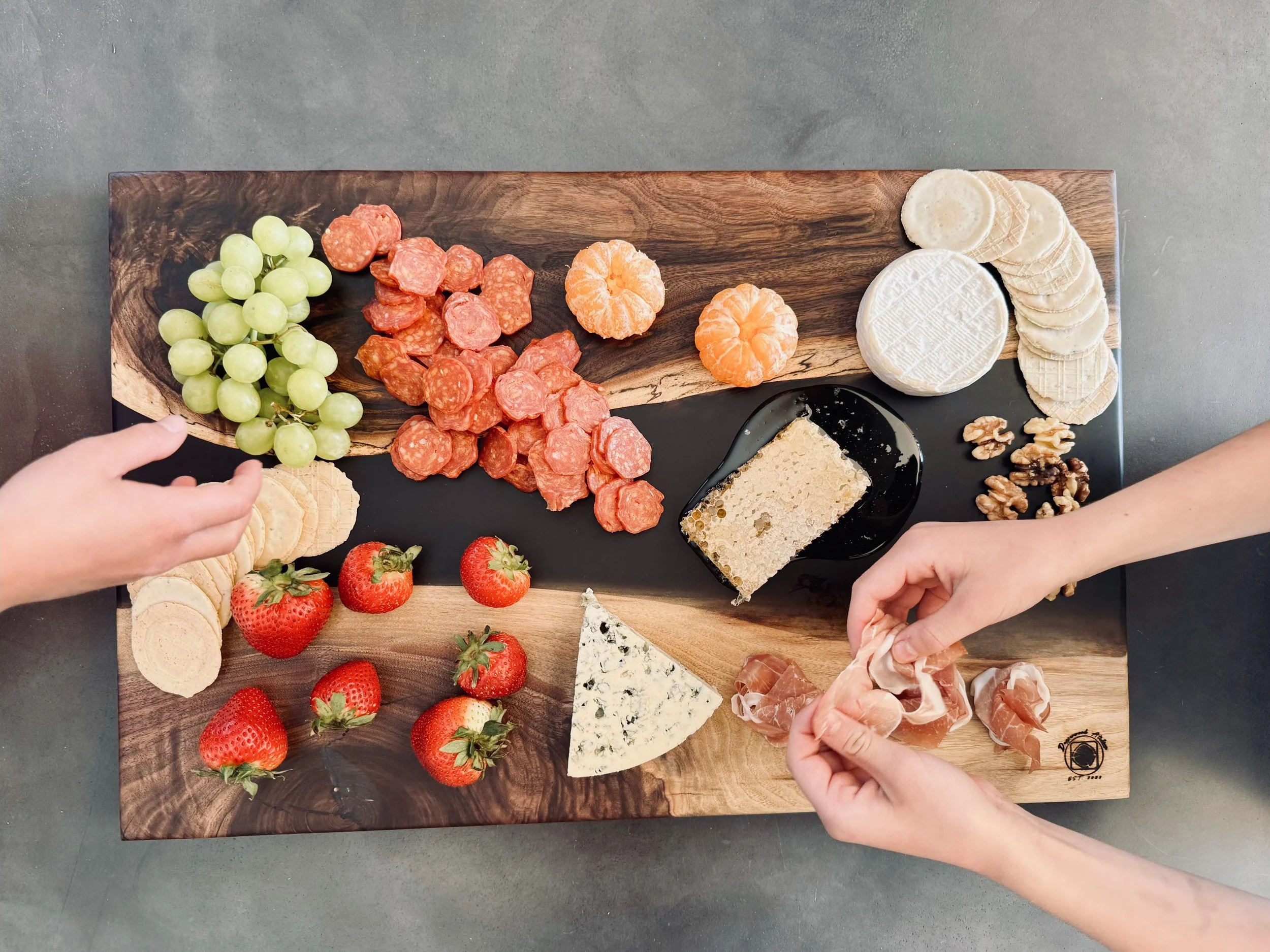 A cheese and charcuterie board featuring grapes, strawberries, various cheeses (including Brie and blue cheese), salami, sliced crackers, walnuts, and two small tangerines on a wooden and black slate platter.