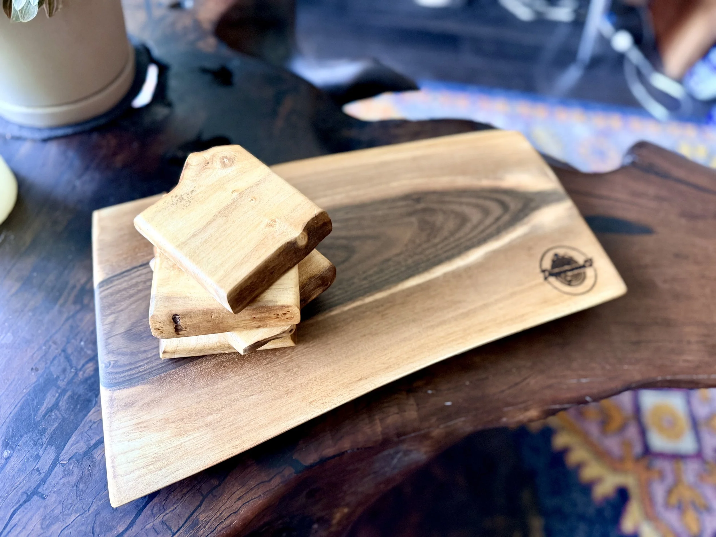 A small wooden sculpture made of three flat rectangular pieces of wood arranged in a stepped formation on a rectangular wooden tray with a logo in the lower right corner. The tray is placed on a dark wooden surface.