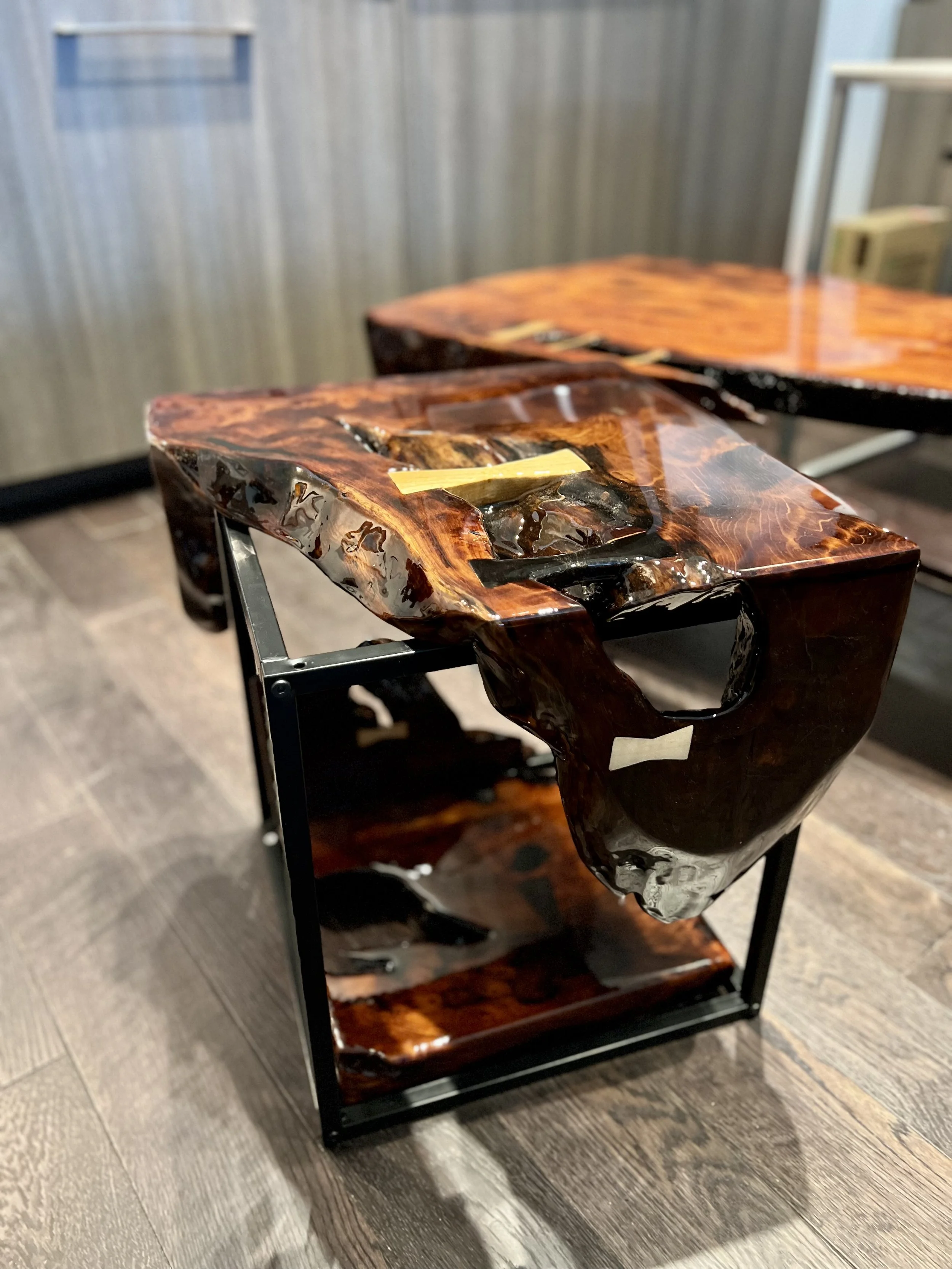 Close-up of a polished, glossy wooden side table with a carved, organic shape and black metal frame, placed on a wooden floor.
