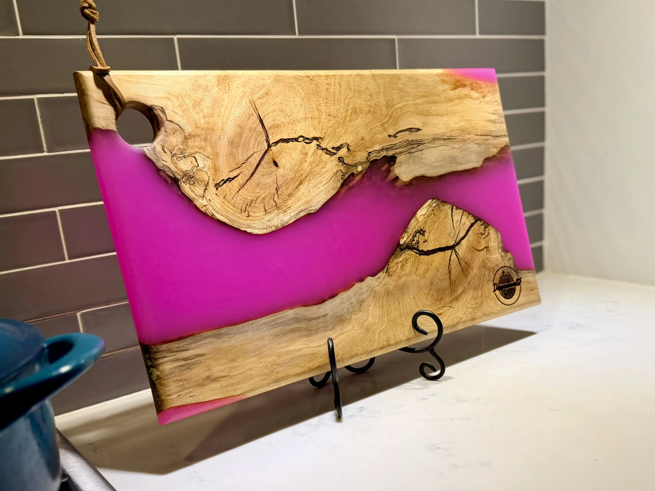 A wooden and pink resin art piece on a black metal stand displayed on a white surface with a gray tiled wall in the background.