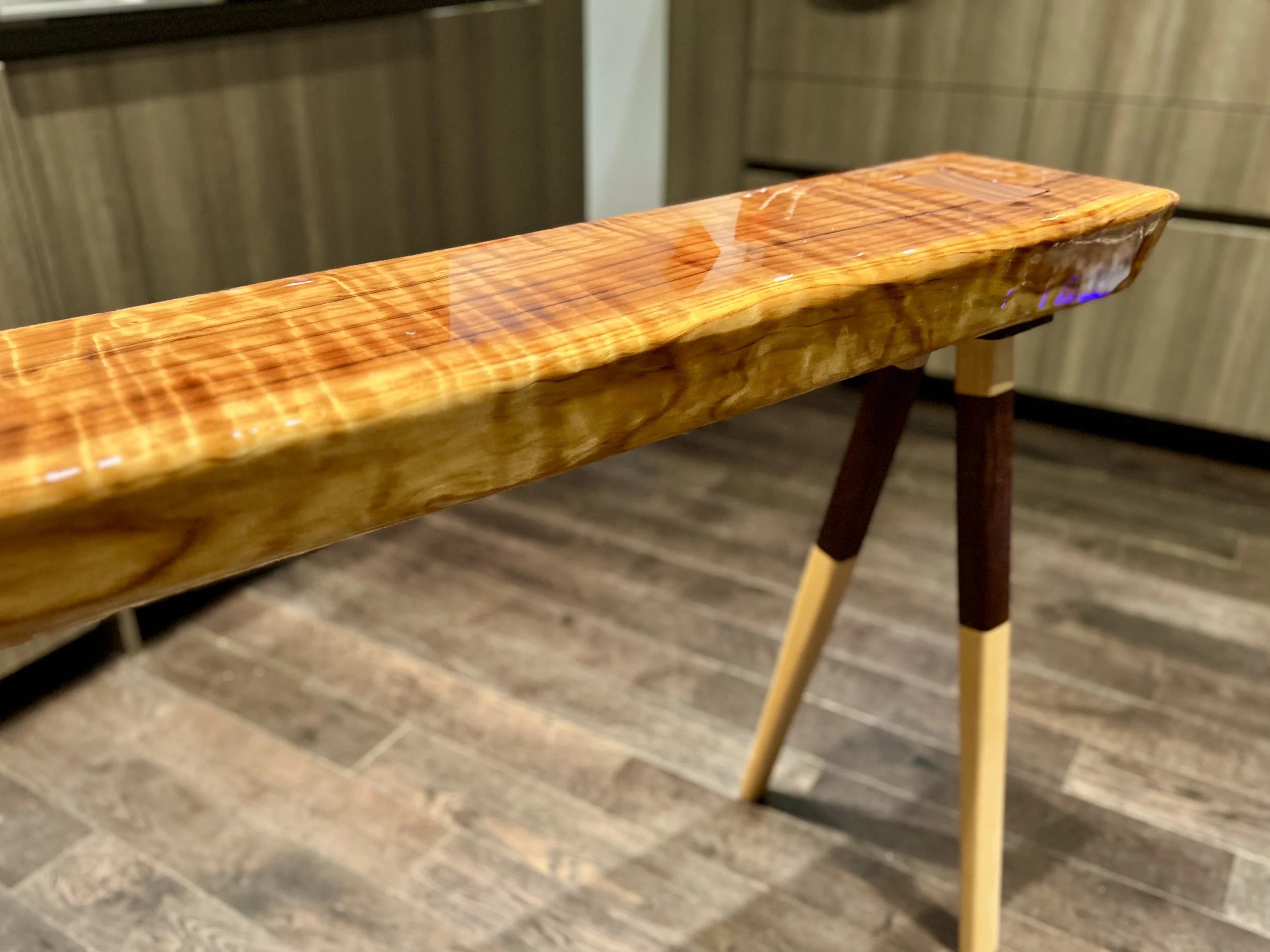 Close-up of a wooden table with a glossy finish, supported by two slender legs, set on a wooden floor in a room with wood-paneled cabinets.