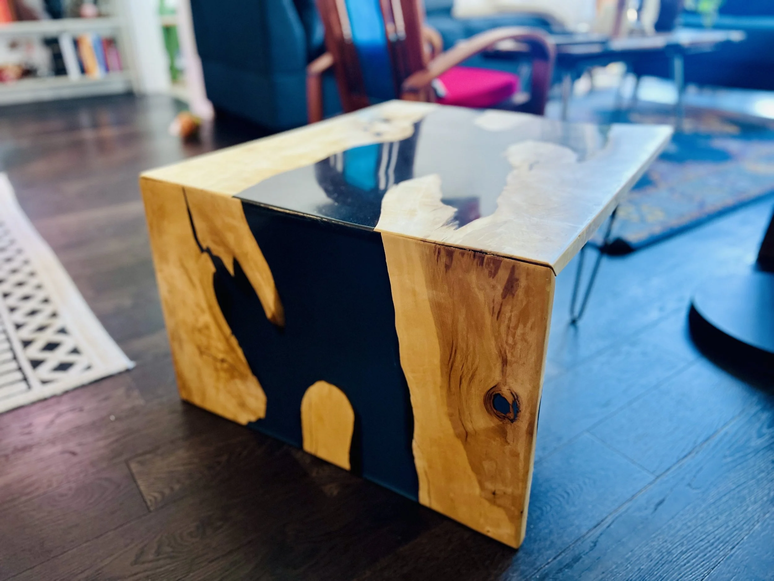 A wooden side table with a black mountain silhouette painted on its side, placed on a dark hardwood floor in a living room.