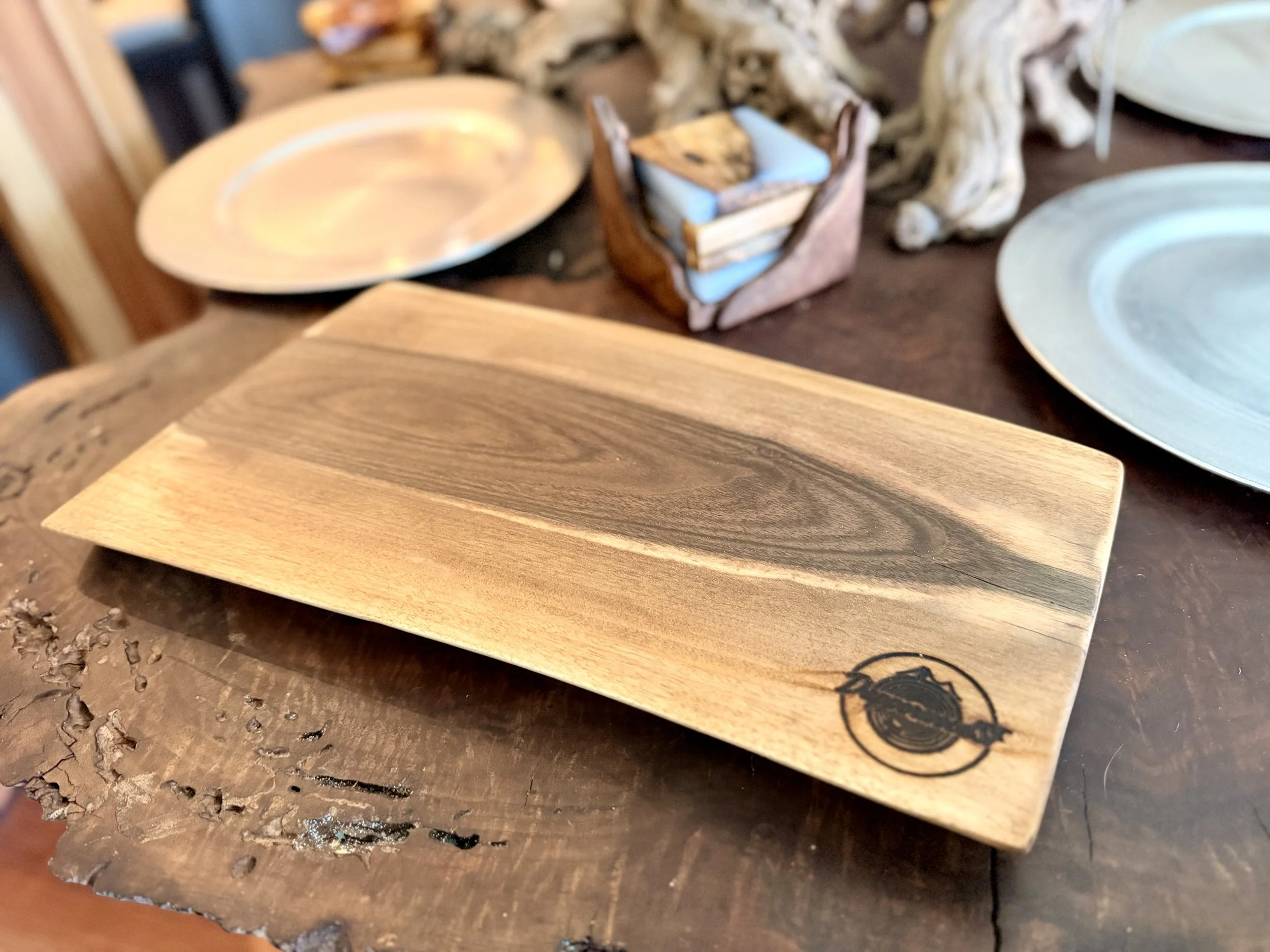 A rectangular wooden serving board with a circular logo in the bottom right corner, placed on a rustic wooden table with ceramic plates and a small wooden container in the background.