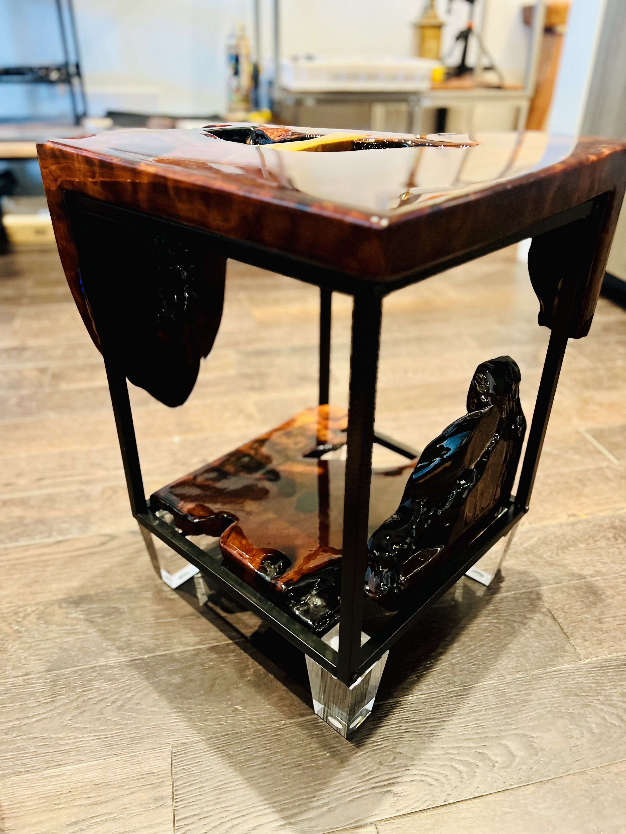 A decorative table with a black metal frame and glossy black and brown resin accents, placed on a wooden floor with a blurred room in the background.