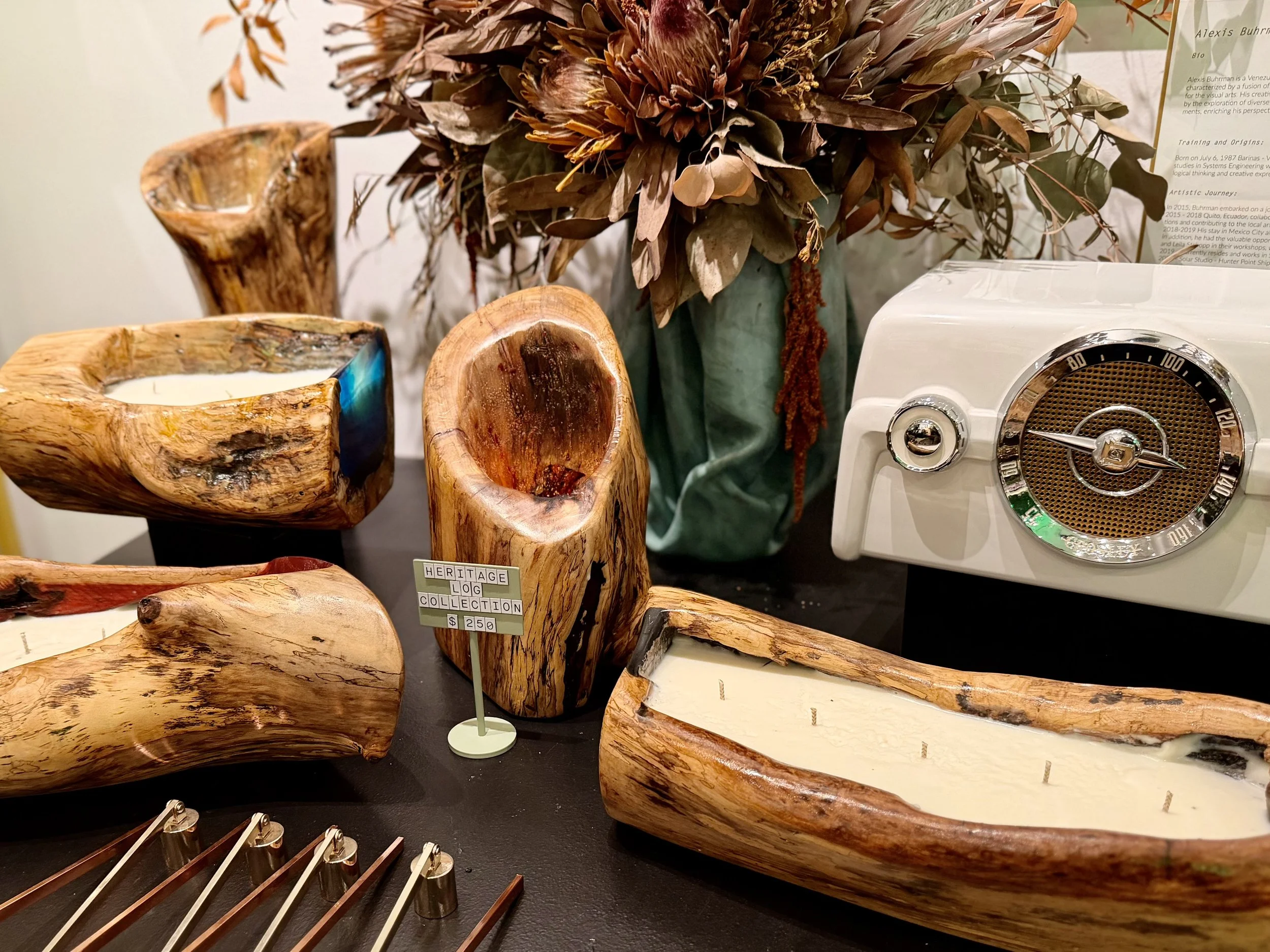 Display of wooden candle holders, a vintage-style clock radio, a vase with dried flowers, and a small sign that reads 'Heritage Log Collection $250'.