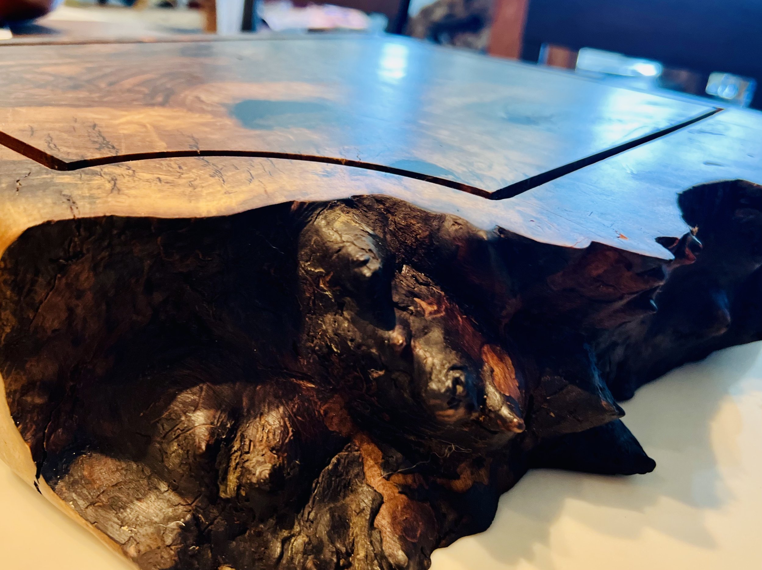 Close-up of a carved wooden table with a rustic design, featuring a bark-like edge. The surface is smooth and polished, with a light natural wood color. The background is slightly blurred.