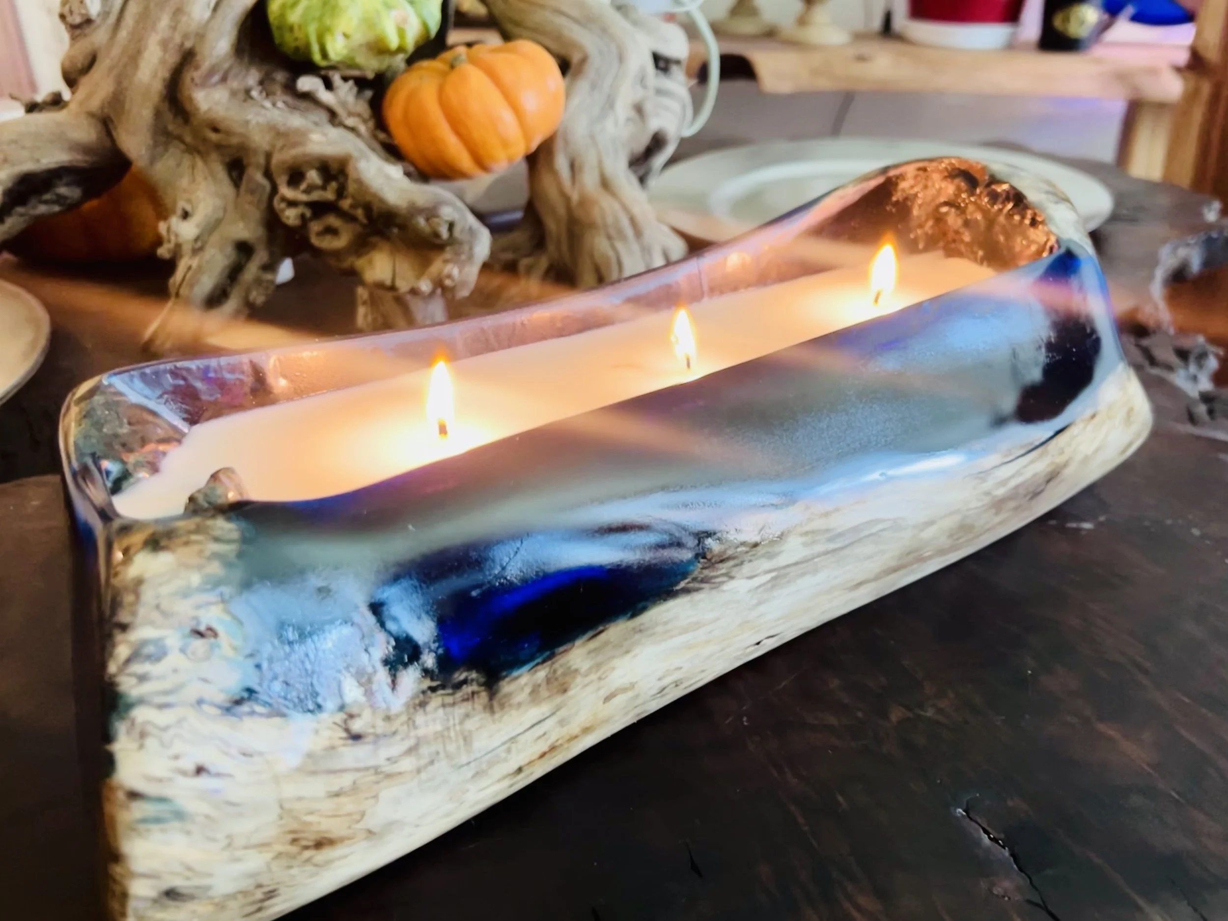 A decorative wooden candle holder with three small flames, positioned on a dark wooden table. In the background, there are small decorative pumpkins and a piece of driftwood with a small pumpkin on top.