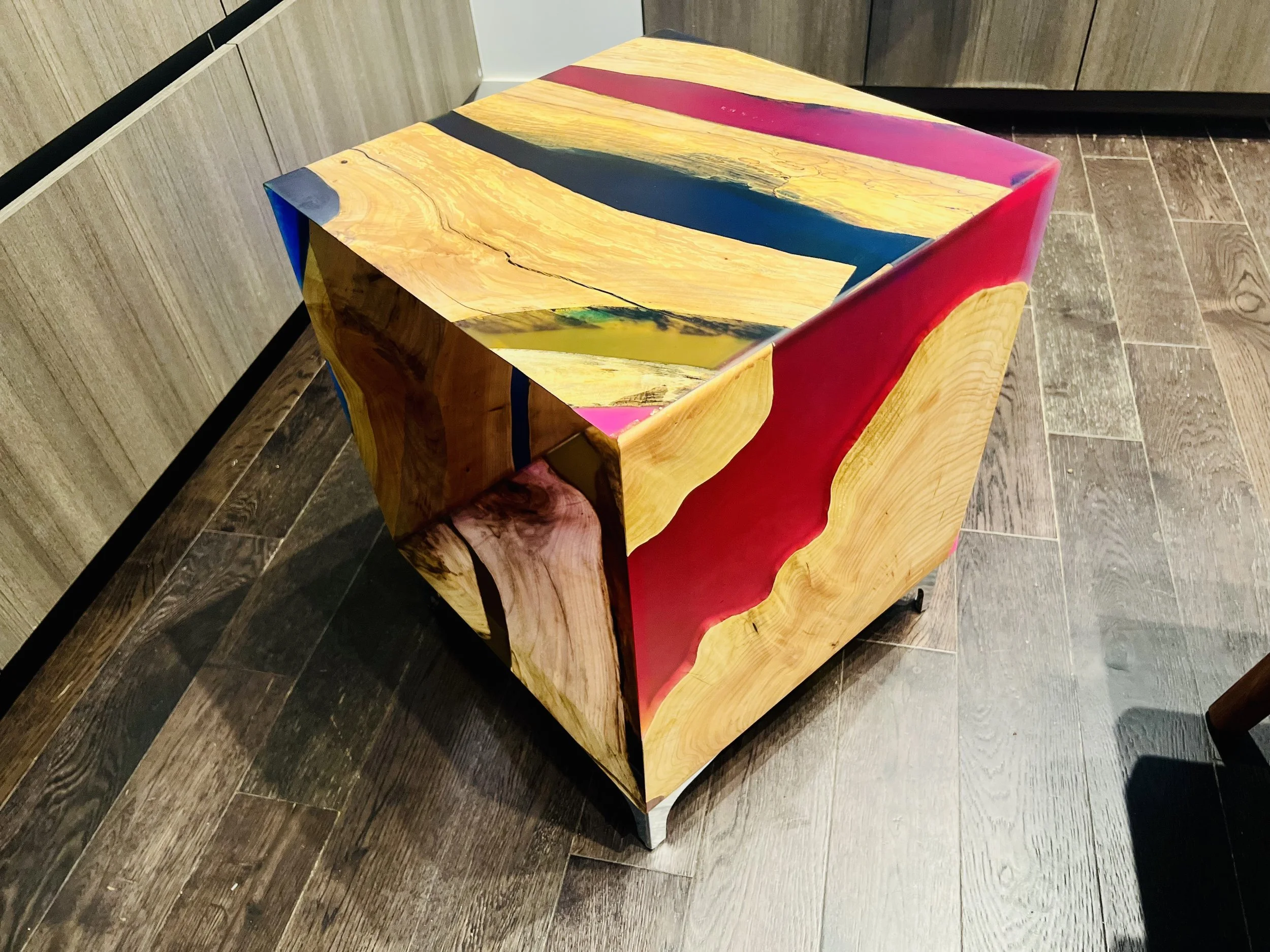 Colorful wooden table with a vivid design featuring natural wood grain and bright red, yellow, blue, and pink accents, placed on a dark wood floor near wooden cabinets.