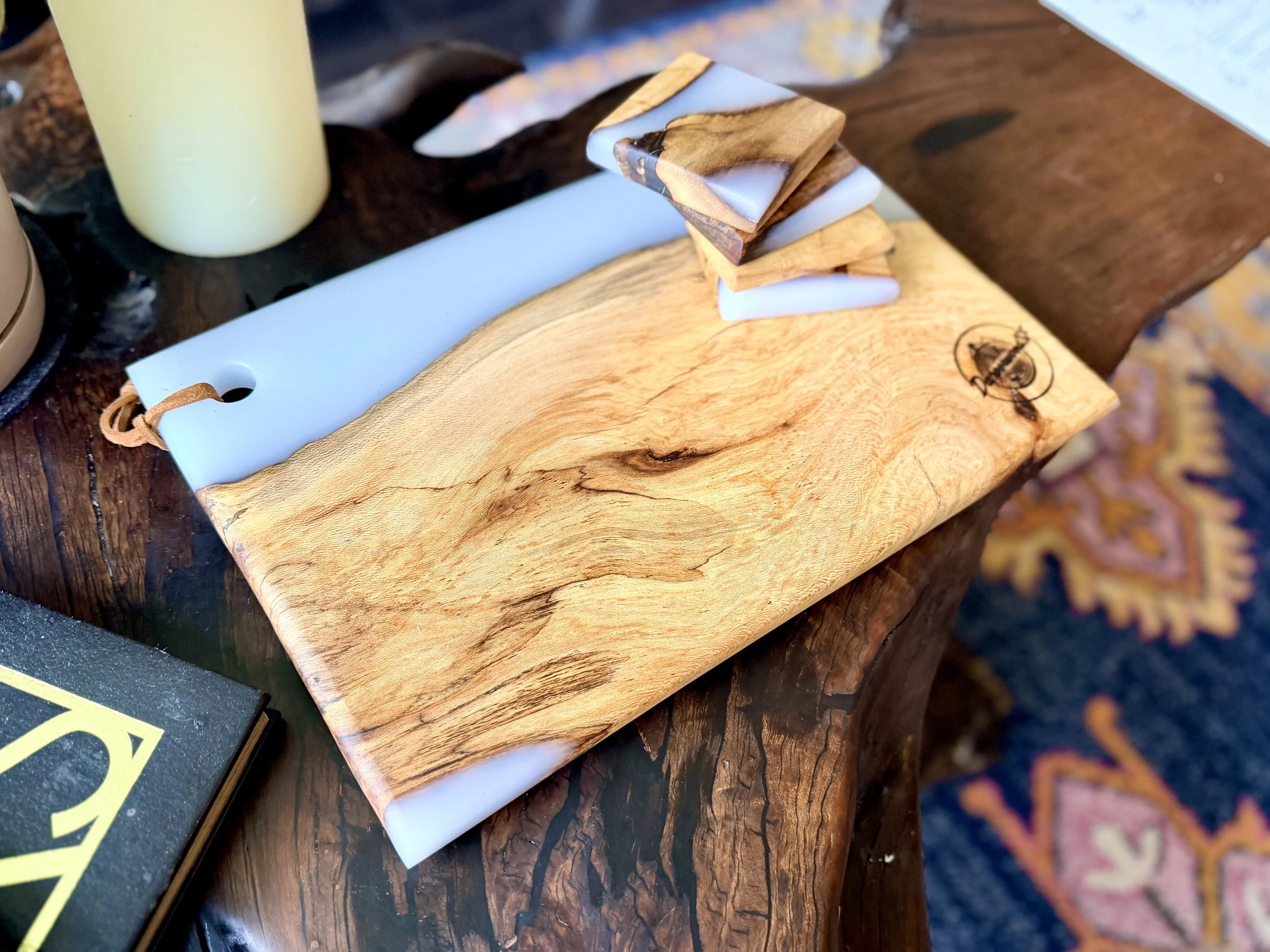 A wooden table with a natural wood finish, holding a large wooden board with a logo, a stack of small wooden and white objects, a yellow candle, and a black book.