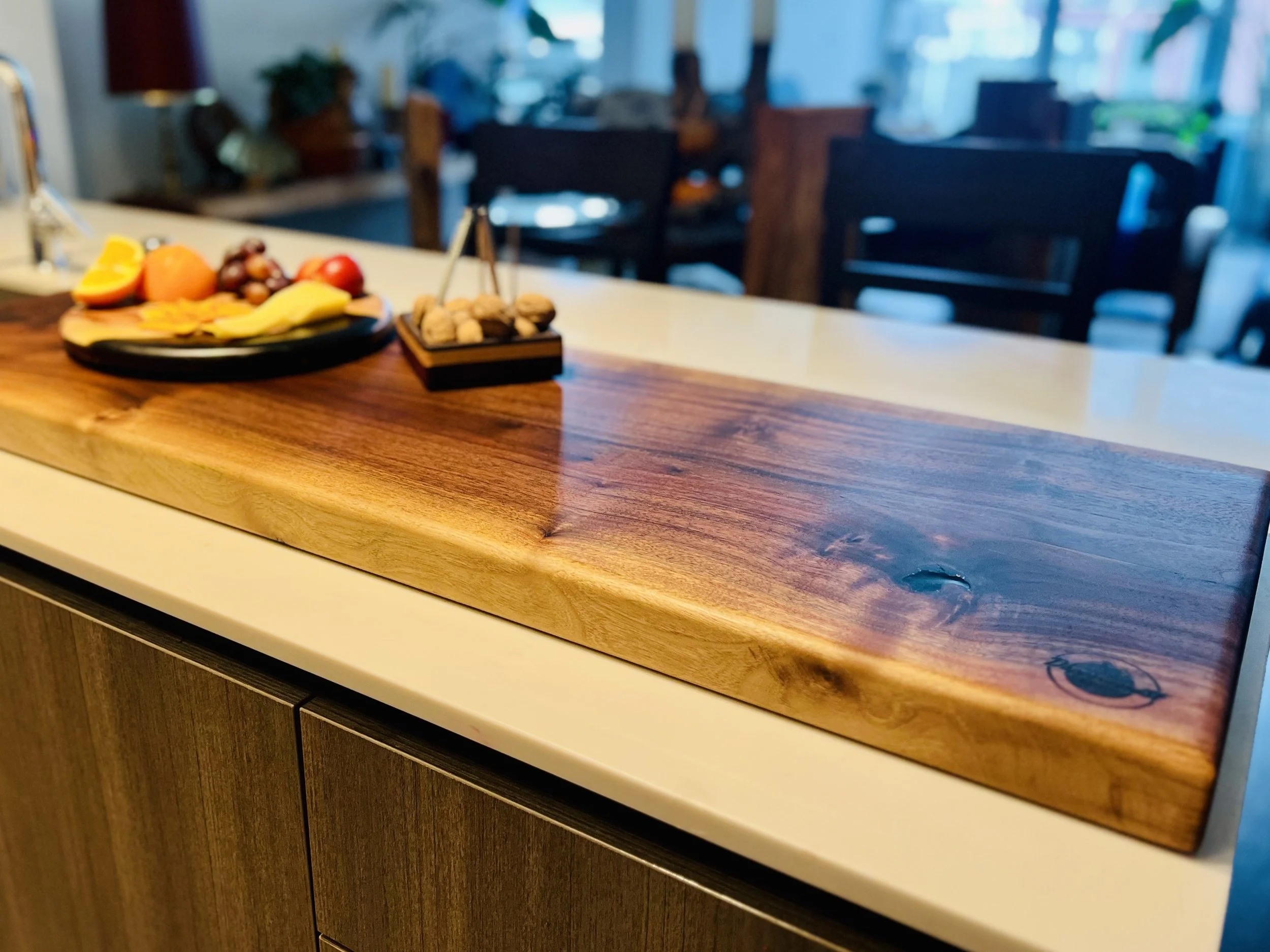 A wooden cutting board on a kitchen counter with a fruit platter and a small bowl of nuts, with a background of a dining room.